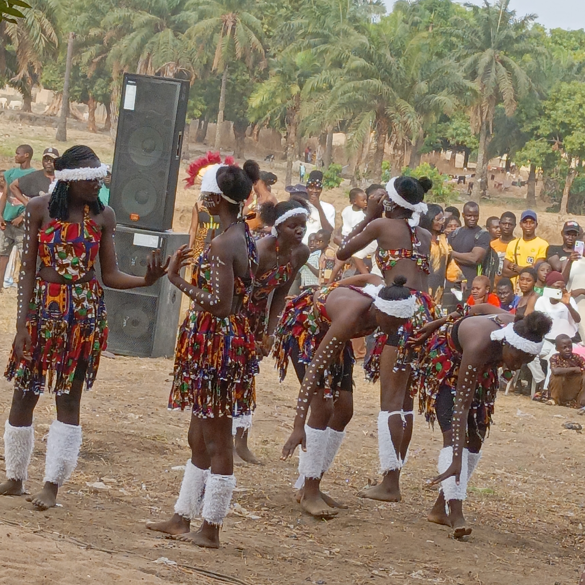 The image captures an outdoor cultural performance in Akwanga, Nigeria. In the foreground, seven young females, identified as dancers, are performing on a light brown dirt surface. They are clad in matching two-piece outfits made of vibrant, multicolored patterned fabric, accented with white headbands and white, fuzzy leg warmers worn on bare legs. Several dancers exhibit white dot patterns painted on their arms and legs. One dancer on the far left stands upright, while others are bent forward at the waist, executing a synchronized movement. To the left of the dancers, two large, black PA speakers are stacked vertically. In the mid-ground and background, a crowd of spectators, predominantly adult males and children, observes the performance. Several audience members are holding mobile phones, appearing to record the event. The background consists of numerous tall palm trees and other green foliage, with distant, indistinct structures visible through the vegetation. The scene is illuminated by natural daylight.