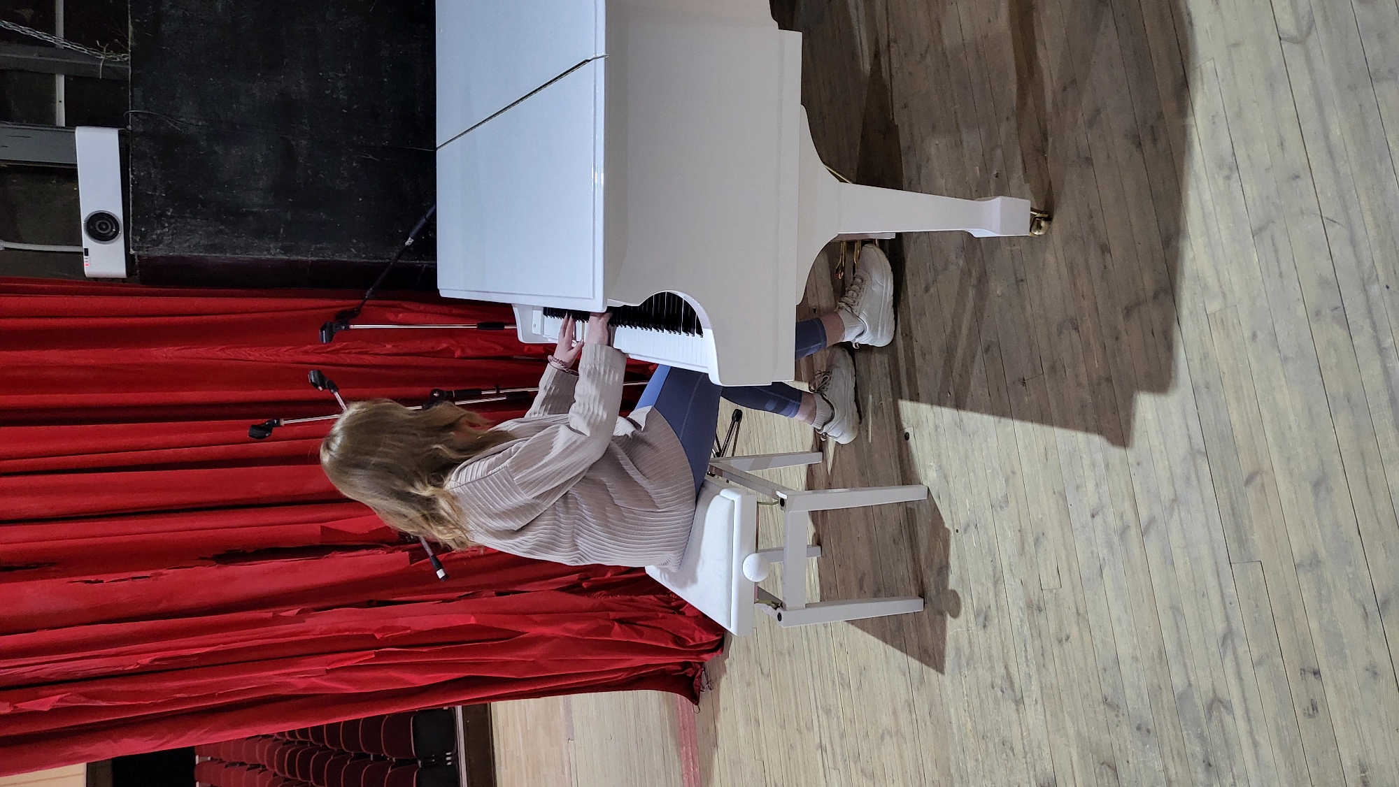 A person with long, light-brown hair is seated at a white grand piano, positioned to play with their hands on the keys. They are wearing a light-colored, possibly beige, striped hoodie, blue leggings, and white sneakers. The piano bench is also white.
The scene appears to be a stage or performance area, characterized by a backdrop of deep red, pleated curtains that extend behind the piano and to the left of the person. Several black microphone stands with metallic poles are visible among the curtains and near the piano. To the far left, a white rectangular piece of equipment, possibly a speaker or monitoring device, with a circular component, rests against a dark wall. Through a gap in the red curtains on the left side of the image, rows of dark theater seats are faintly visible, suggesting an auditorium or theater setting. The floor of the stage is made of light-colored wooden planks, with shadows cast across it, indicating indoor artificial lighting. No text is visible in the image.