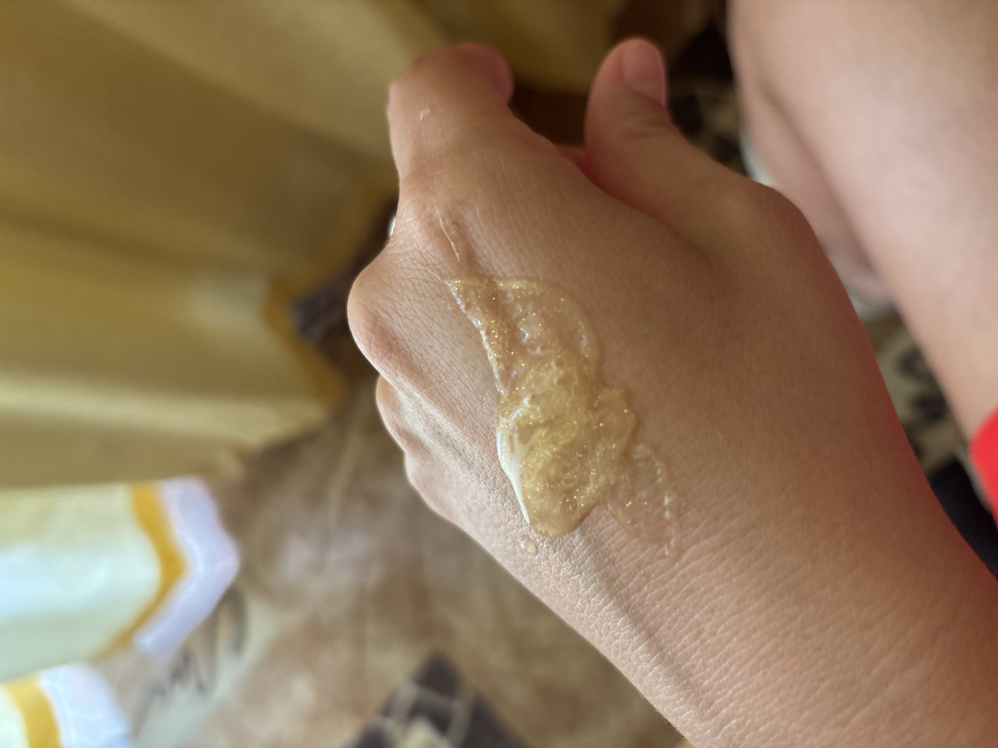 A close-up shot shows a person's hand with a dollop of shimmering, golden gel on the back. The gel appears somewhat viscous and contains fine glitter particles. The skin of the hand is visible with texture and pores. The background is softly blurred, showing elements of an indoor environment with what appears to be yellow curtains and a patterned surface, possibly a rug or blanket. The image does not contain any elements that would allow for identification of a specific location, including Krasnaya Polyana, Russia.