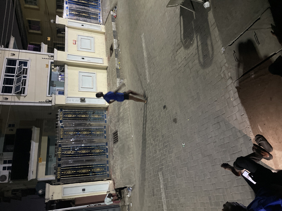 An image captured at night depicts a street scene. A person in a blue shirt and dark shorts appears to be running or walking quickly. Adjacent to the street are buildings with decorative gates. At the lower right of the frame, another person is visible, seated and using a smartphone. The street is paved with bricks or tiles. Shadows are cast from an unseen light source.
