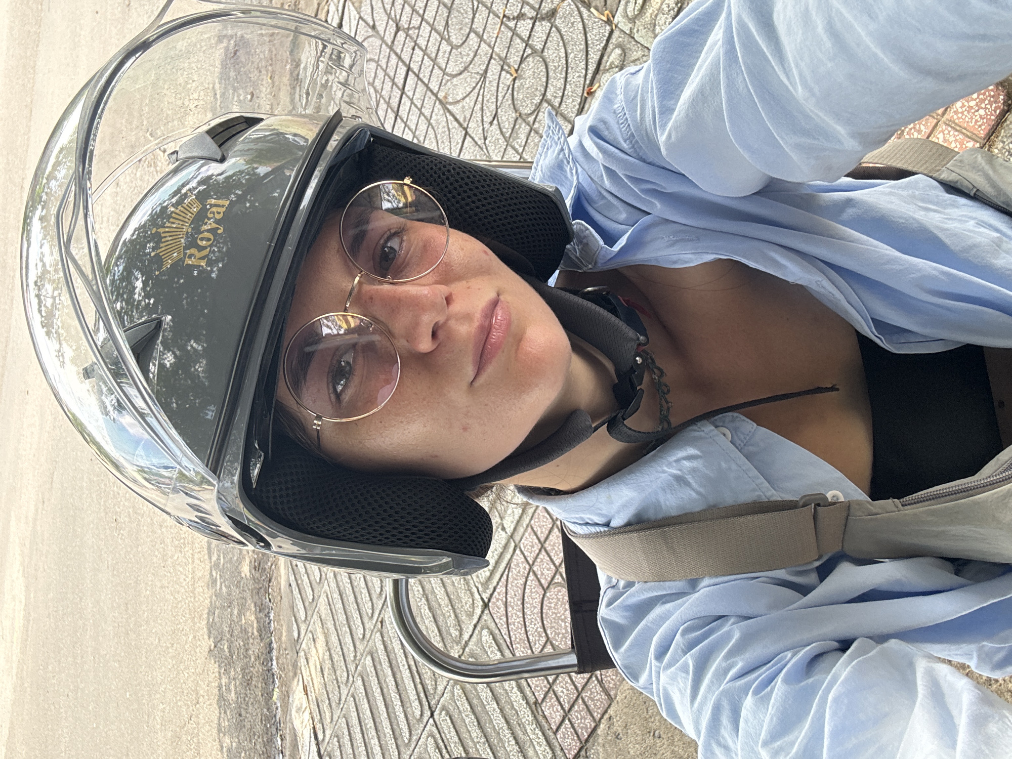 The image depicts an individual, identified as an adult female, in a selfie taken outdoors in Phu Thuy Ward, Vietnam. She is wearing a dark grey open-face helmet with a clear visor currently in the upright position. A gold emblem featuring a crown and the word "Royal" is affixed to the front of the helmet. Round, gold-rimmed spectacles are worn over her eyes, and freckles are visible on her nose and cheeks. Her attire consists of a light blue long-sleeved button-up shirt, unbuttoned over a black undergarment. A light grey strap, consistent with a shoulder bag, is visible across her right shoulder. A dark tattoo is partially visible on her lower neck/upper chest area beneath the helmet strap. Behind the individual, a curved silver metallic bar, possibly a seat back or vehicle handle, is present. The immediate ground surface features patterned paving stones with red and light-colored geometric squares. Further back, a light-toned asphalt street or pathway is visible. The scene is illuminated by natural daylight.