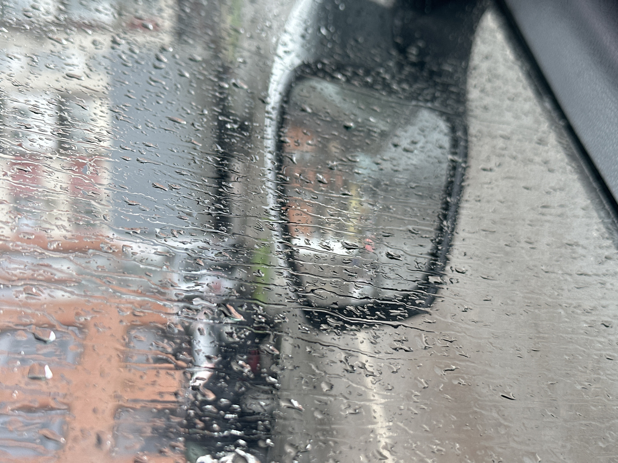 The image shows a rain-covered car window with a side mirror visible. Through the window, blurred buildings, possibly brick, are seen, reflecting the wet conditions of a city street. The raindrops streak across the glass, distorting the view outside. The interior of the car includes a partial view of a dark gray or black panel, likely part of the car's frame. The image does not allow for determination of a specific location in Rostov-on-Don, Russia, nor does it contain enough detail to determine information about people, interactions, or actions.