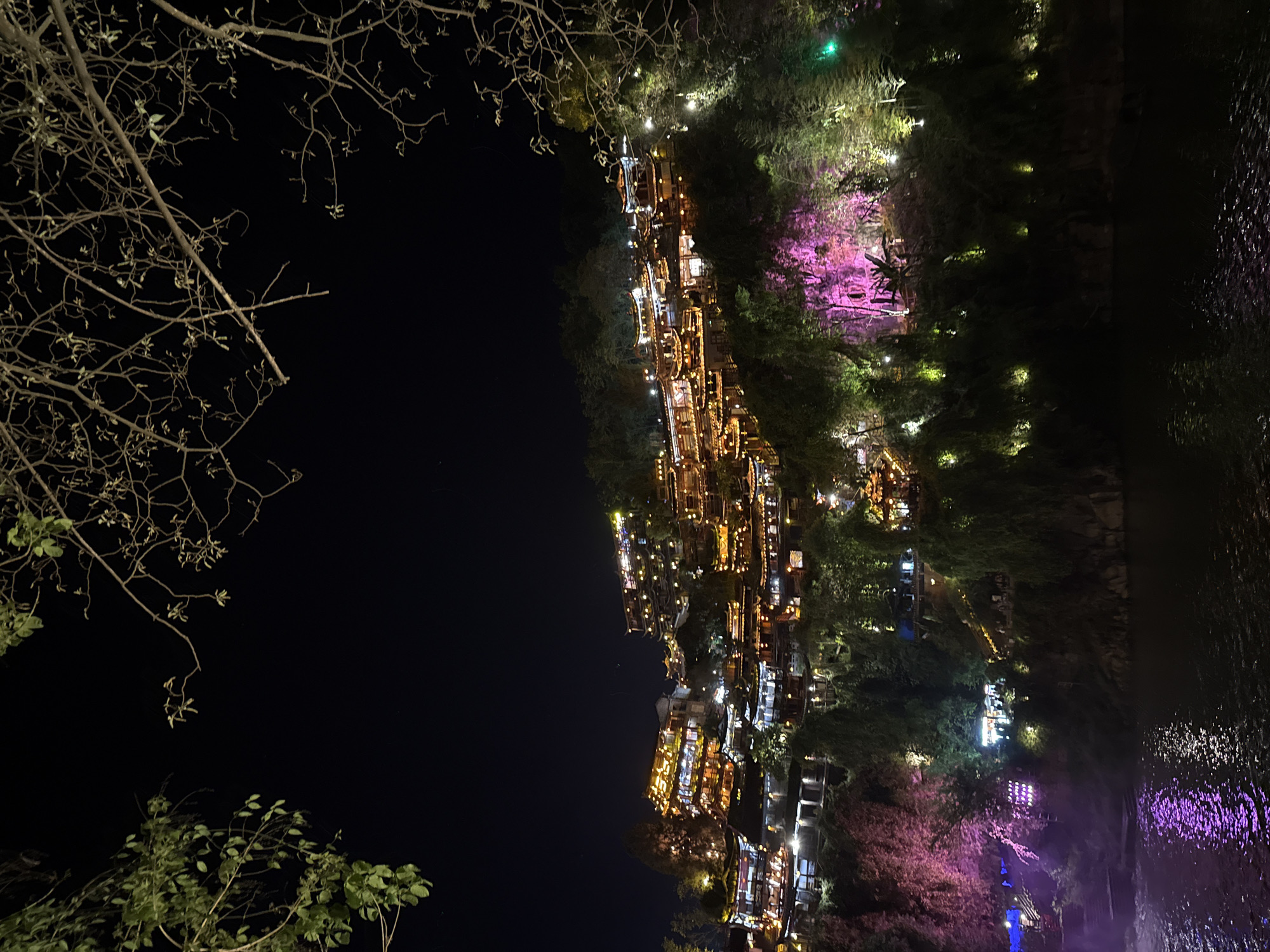 The image shows Furong Ancient Town in Hunan Province, China, at night. The town is illuminated with warm, orange lights, creating a warm glow. The lights reflect off the water. The scene is framed by the silhouettes of trees in the foreground. The buildings appear traditional with multiple levels. Some vegetation around the town is lit with green and pink lights. A body of water, a river, is on the right.