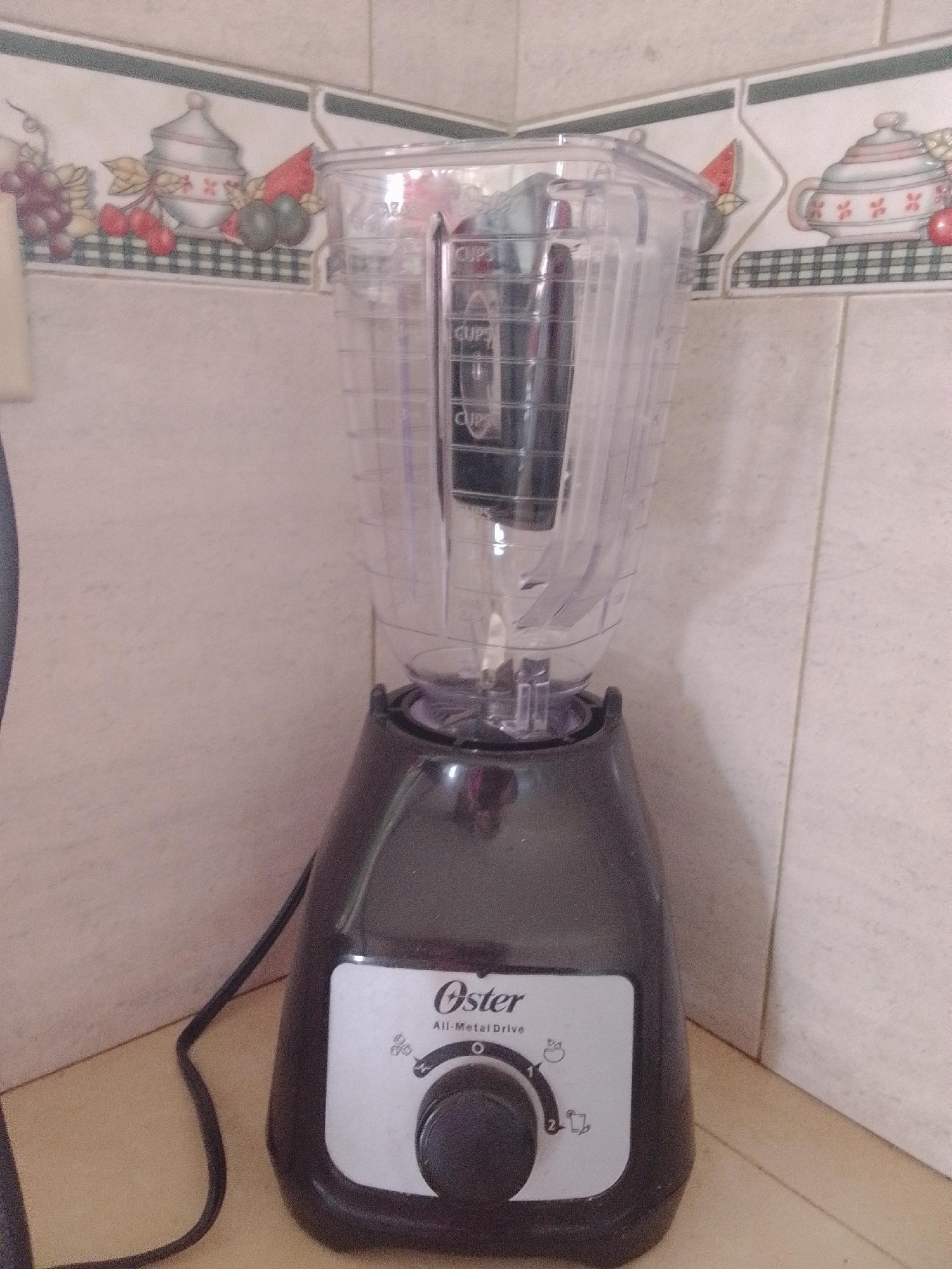 In La Tagua, Venezuela, an Oster All-Metal Drive blender sits on a countertop against a tiled backsplash. The blender is black with a clear plastic pitcher. The pitcher has measurement markings visible. The control panel on the blender's base displays various speed settings indicated by icons. To the left of the blender, a black power cord is visible. The backsplash features a decorative border with images of fruit and pottery.