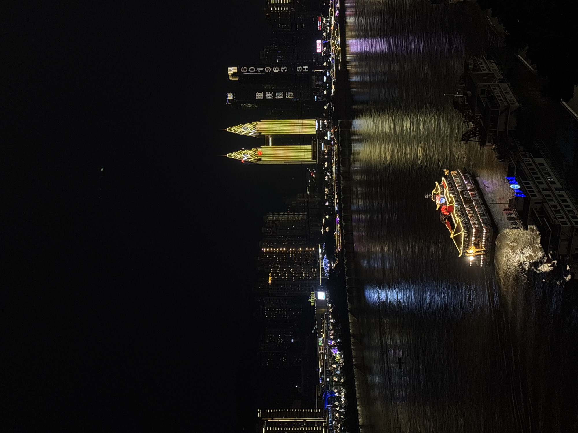 Here is a description of the image:
This is a night scene along a body of water, likely a river, in Yuzhong District, China. The lights of a cityscape are visible on the opposite bank, with tall buildings illuminated with various colors and patterns. One building displays the text "SH 3993106", and another with Chinese characters. The water reflects these city lights, creating shimmering patterns on the surface. A brightly lit boat moves along the river, also casting a glow onto the water. Several other stationary boats are docked along the shoreline. The overall lighting is dim due to the darkness of the night, and the city lights provide the primary source of illumination.