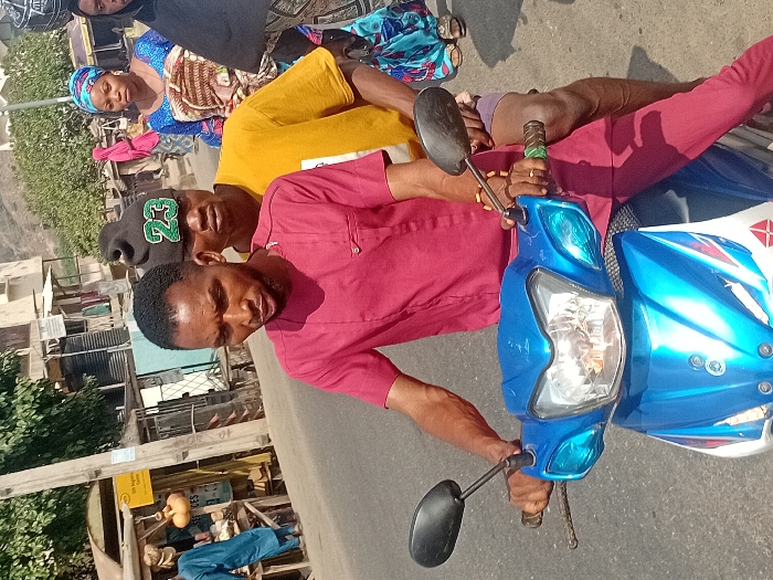 A man in a red shirt drives a blue and white motorcycle. Another man, wearing a yellow shirt and a black baseball cap, sits behind him. A woman in a patterned blue top sits behind the second man, facing forward. They are on a paved road, with buildings and other people visible in the background. A streetlight stands beside a building to the left of the frame.