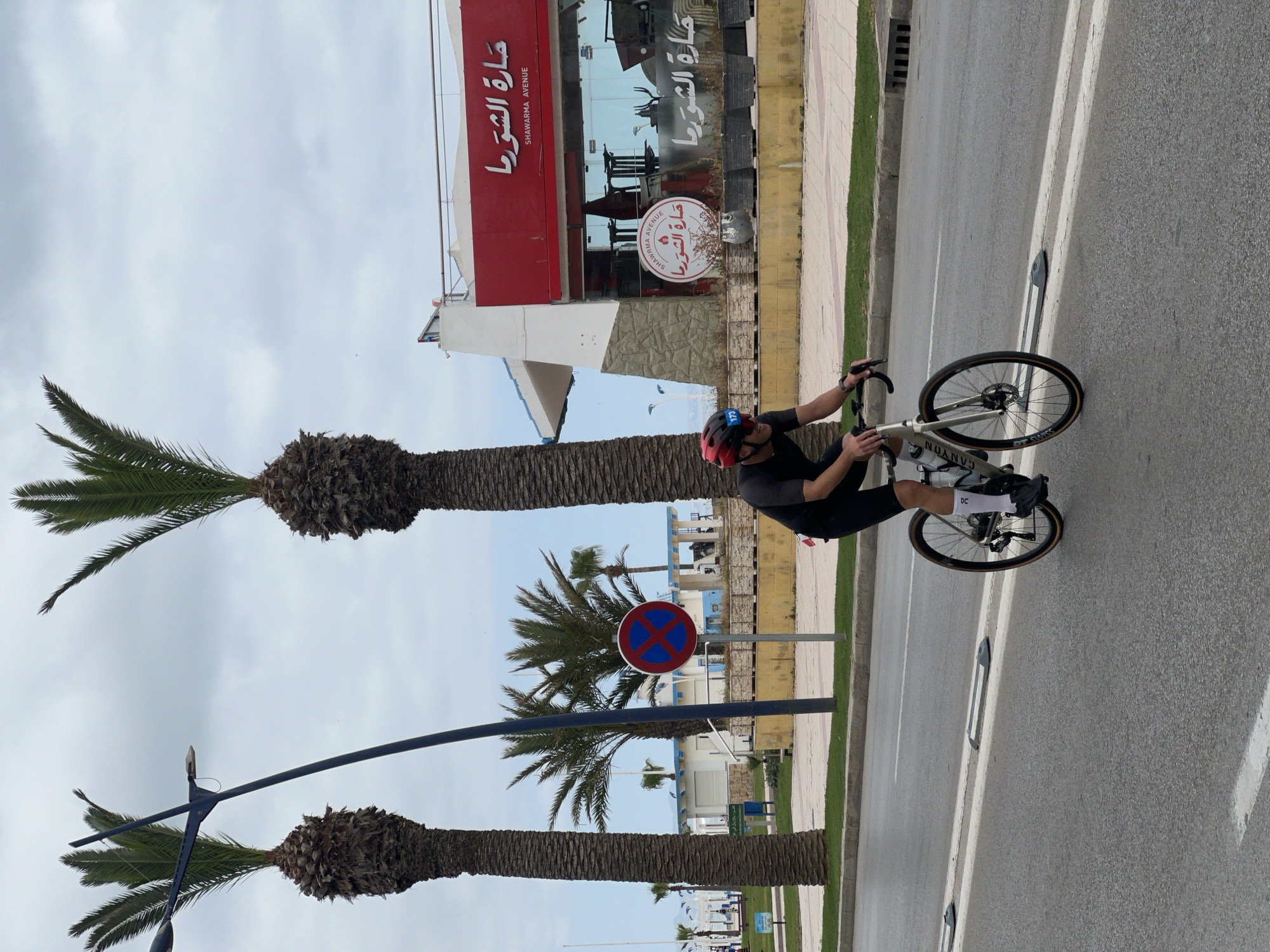 A cyclist wearing a red helmet and dark clothing rides a white Canyon bicycle on a paved road. The location is identifiable as M'Diq, Morocco, due to the presence of a building with Arabic script reading "Shwarma Avenue" and "حارة الشورما". Palm trees line the street. A blue and red no-parking sign is visible in the background. The sky is overcast.