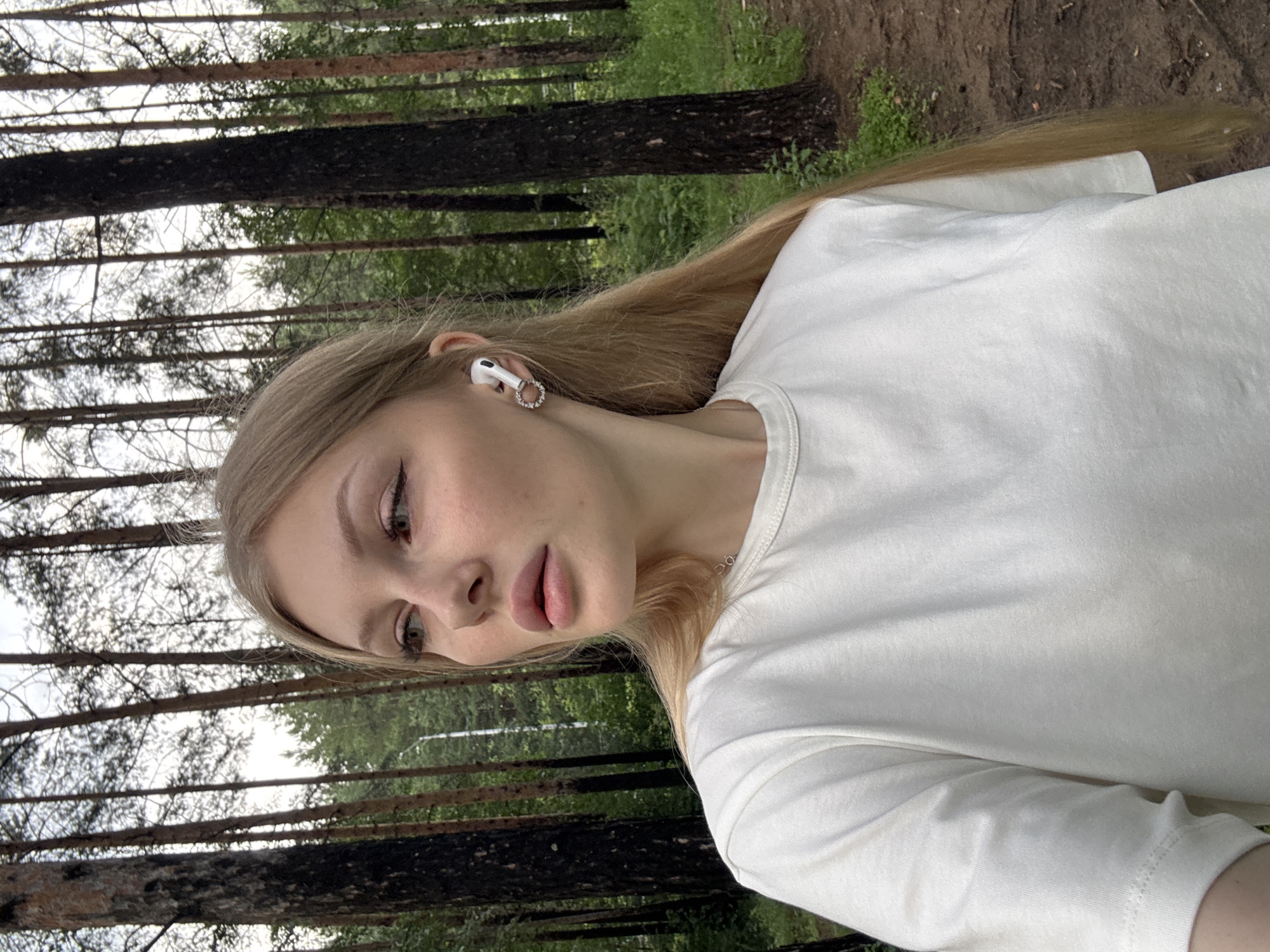 A young woman with long blonde hair, wearing a white t-shirt and white earbuds, is seen from the chest up, outdoors in a forest setting. She has her head turned to her left, and her gaze is directed away from the camera. The background consists of tall pine trees and greenery, indicating a natural, wooded environment. The lighting suggests it is daytime, possibly overcast. The location is not specified in the image itself, therefore a specific location in the city of Perm, Russia, cannot be determined.