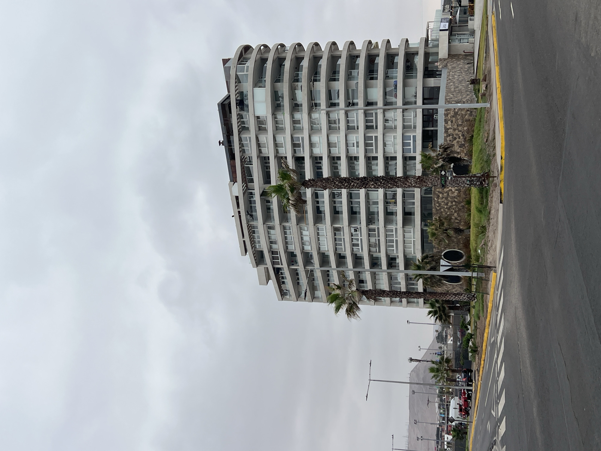 The image shows a tall, modern building in Arica, Chile. The building is white with a curved design and many balconies. Palm trees line the sidewalk in front of the building. A road runs along the side of the building, with cars and pedestrians visible in the distance. The sky is overcast.
