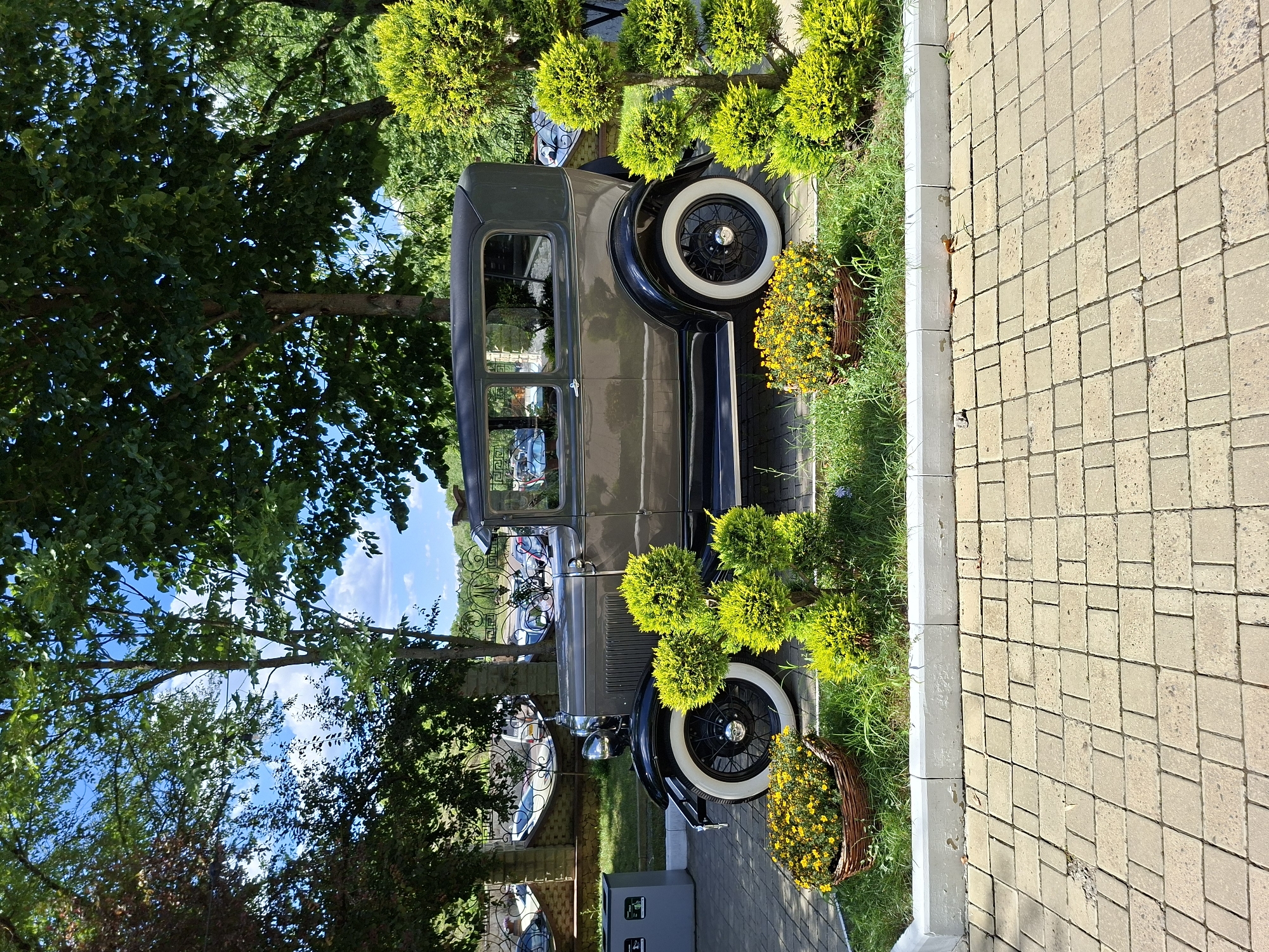 The image shows a vintage, dark grey two-door sedan parked on a paved walkway in front of a grassy area with decorative bushes and flowers. The car appears to be a Ford Model A, characterized by its spoked wheels with white sidewalls and a dark-colored canvas roof. The background reveals trees, a decorative metal fence, and other parked vehicles, suggesting an outdoor public space, possibly a park or event venue. The area is well-lit, indicating it is daytime. The paved walkway is made of rectangular light-colored stones. The grassy area features neatly trimmed bushes and a woven basket filled with yellow flowers. This scene is located in Milestii Mici, Moldova, as indicated by the context provided. There are no people visible in the image.