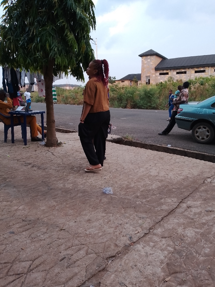 The image depicts an outdoor scene in Umueshi, Nigeria. A woman with red braids stands on a concrete sidewalk, facing away from the camera. She wears a brown shirt and black pants. To the left, a man sits at a table under a tree, appearing to work on a laptop. Laundry hangs on a line near the tree. In the background, across a road lined with green vegetation, a building with a distinctive tower structure is visible. To the right, a man leans against a teal car, holding a child, and another child stands nearby. Litter is scattered on the sidewalk.