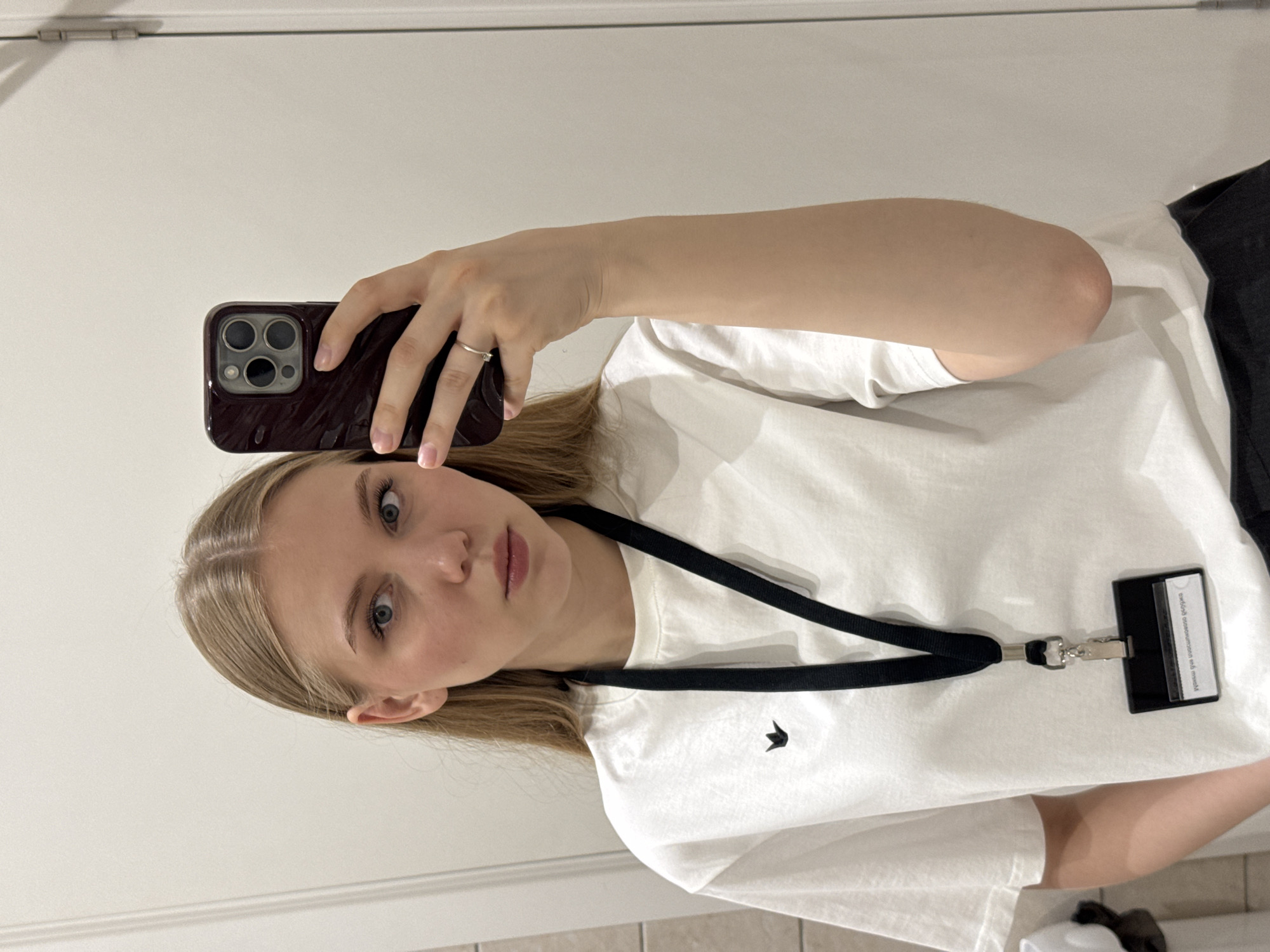 This image shows a young adult female capturing a mirror selfie indoors. The subject has straight, light blonde hair parted in the middle, blue eyes, and a neutral expression. She is wearing a white, short-sleeved, crew-neck t-shirt adorned with a small, dark, crown-like emblem on the left chest. A black lanyard is worn around her neck, from which a black badge holder hangs, partially revealing an ID card with inverted Cyrillic text. Her right hand, wearing a plain silver ring on the ring finger, holds a dark-colored smartphone with a prominent multi-lens camera array, pointing towards her reflection. The visible background is a plain, light-colored wall, with a door hinge discernible on the upper right side. The image contains no visual elements to corroborate a location in the city of Perm, Russia.