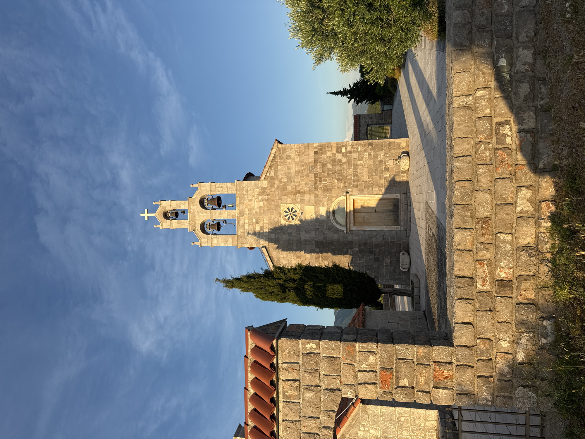 The image depicts a stone church in Kovačko Polje, Montenegro, bathed in the warm light of late afternoon or early morning. The sky is a clear, bright blue with wispy white clouds. In the foreground, a low stone wall with a red-tiled structure on its left separates the viewer from the church complex. The wall is constructed of rough-hewn gray stones with visible mortar. A metal gate is partially visible on the left side of the wall.
Dominating the center of the image is the stone church, constructed from large, rectangular gray stones. It features a prominent bell tower with three bells and a cross at its apex. The bell tower has arched openings and decorative stone carvings. The main facade of the church has a large wooden door with intricate carvings and a circular decorative element above it. A tall, slender cypress tree stands to the left of the church, casting a long shadow across the paved courtyard. To the right of the church, an olive tree with lush green foliage adds to the Mediterranean feel.
In the background, through an arched gateway, more of the church complex is visible, along with hints of rolling hills or mountains under the blue sky. The overall impression is one of tranquility and rustic beauty, characteristic of a Montenegrin village setting.