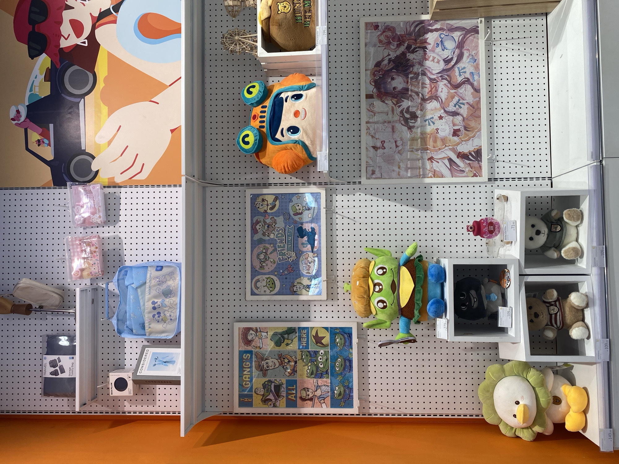 The image depicts a retail display for plush toys and merchandise, likely within a department store or specialty shop. The display features a pegboard wall with shelves, showcasing a variety of items. On the top shelf, there are travel bags and small pouches with cartoon characters. The middle shelf displays a "Toy Story" themed poster and a large plush character, along with a cartoonish character head pillow. Below this, more plush toys are arranged on cube shelves. The bottom shelf and foreground show additional plush animals, including a duck and bears, and a framed anime-style print. The overall impression is a colorful and whimsical collection of goods targeted at a younger audience or fans of popular franchises. The context of North Jakarta, Indonesia, suggests this scene is within a shopping mall or retail area in that region.