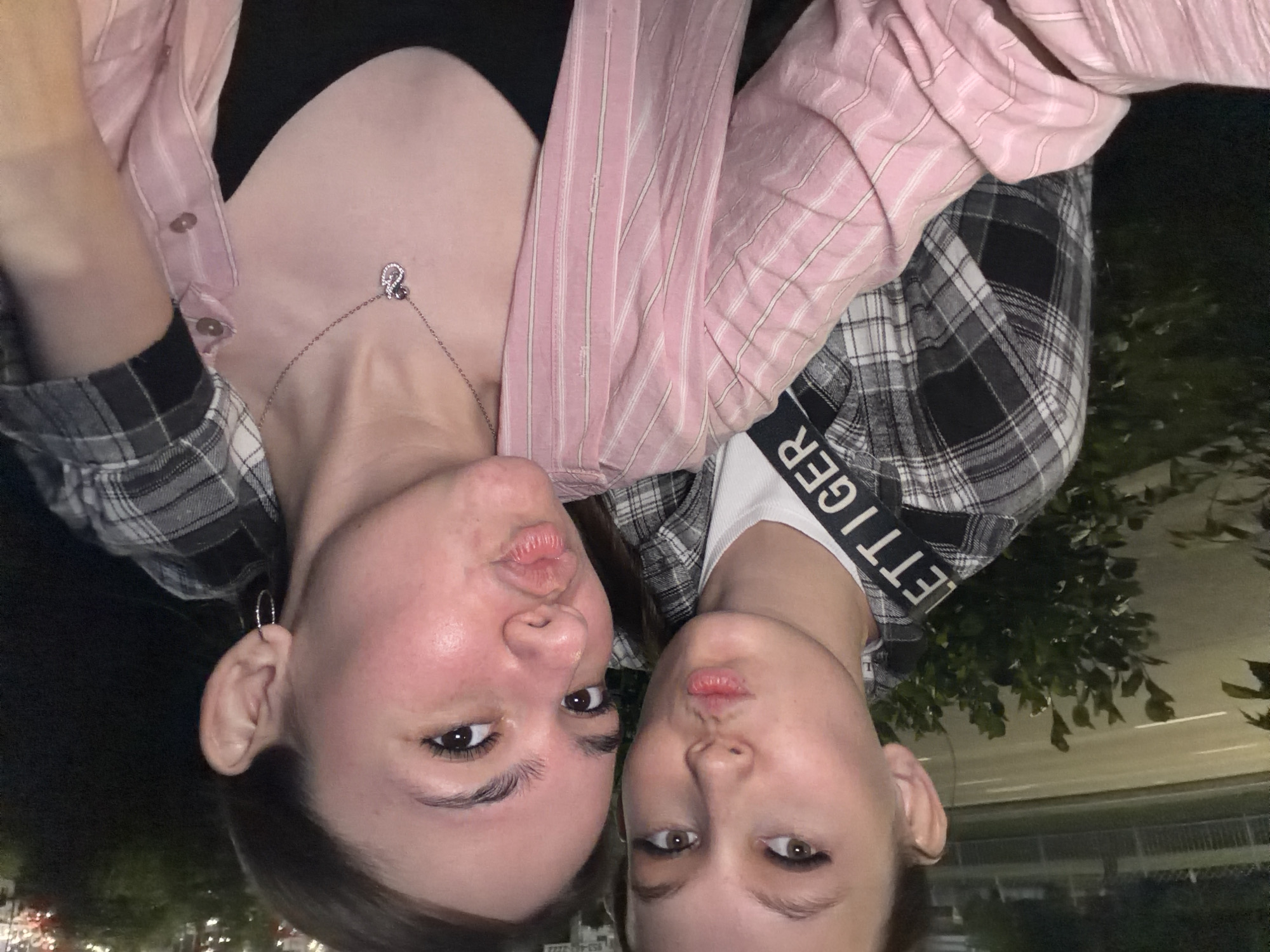 Two young women are taking a selfie outdoors at night. The woman in the foreground is wearing a pink striped shirt over a black top and a necklace. The woman behind her is wearing a plaid shirt. Both women are pouting their lips. The background shows a street with cars and trees. The location is the city of Naberezhnye Chelny, Russia.