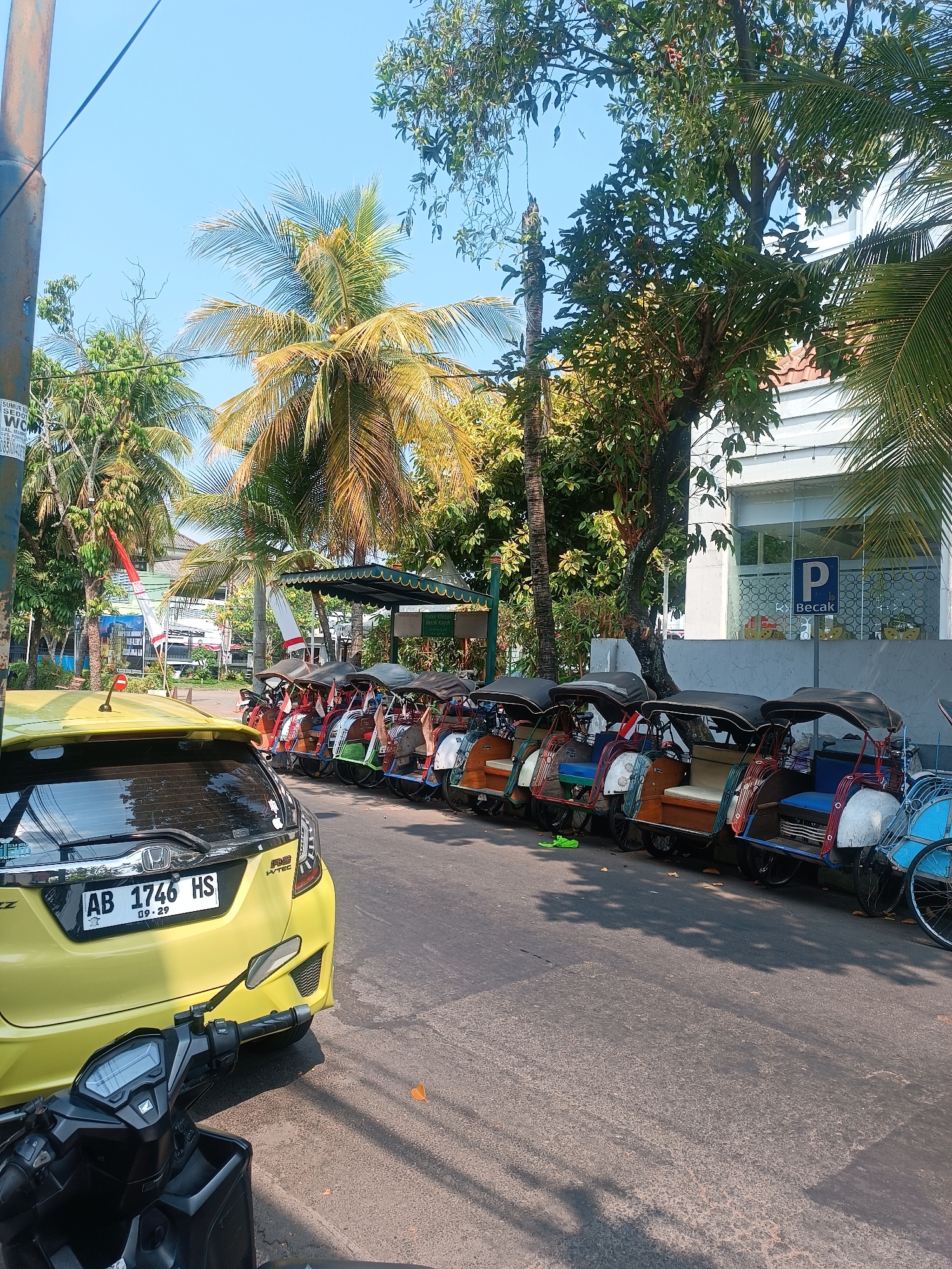 This image depicts a street scene in Yogyakarta, Indonesia, likely near a tourist area or transport hub. In the foreground, the rear of a bright yellow car is visible, with its license plate reading "AB 1746 HS". To the left, a motorcycle is partially visible. The main focus of the image is a row of colorful becaks (three-wheeled cycle rickshaws) parked along the side of the road. These becaks are of various colors, including orange, blue, and red, and appear to be waiting for passengers. Behind the becaks, a building with white siding and glass windows is visible. A blue parking sign with a white "P" and the word "Becak" is prominently displayed on the building's facade. Palm trees and other lush greenery surround the scene, suggesting a tropical environment. The sky is clear and blue, indicating a sunny day. The overall impression is one of a typical street scene in a vibrant Indonesian city, with traditional transportation methods coexisting with modern vehicles and infrastructure.