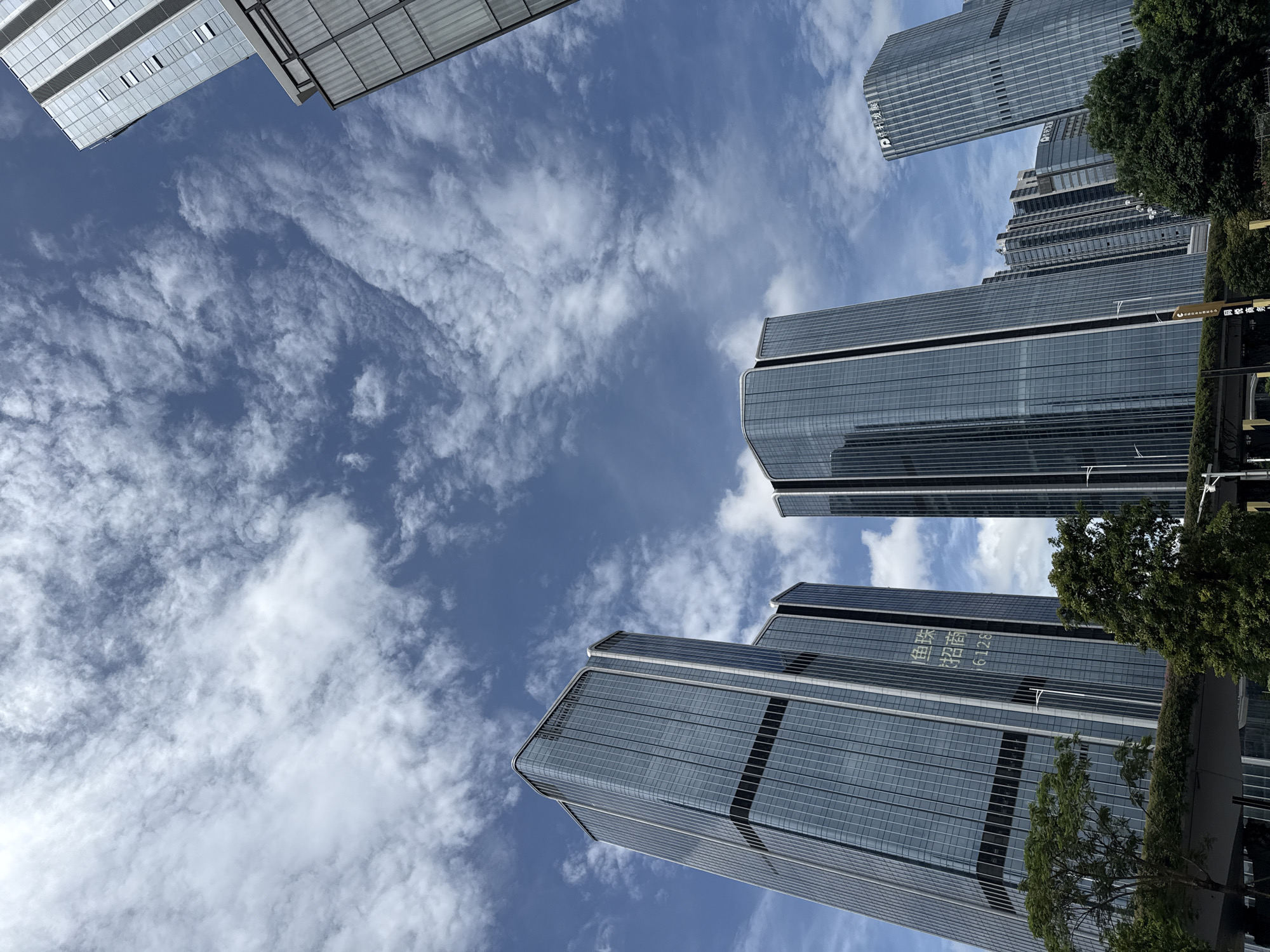 This is a low-angle, wide-shot photograph of modern skyscrapers in the Huangpu District, China, under a partly cloudy blue sky. The buildings are tall, with reflective glass facades, and some feature distinctive architectural elements like rounded corners and vertical accents. In the foreground, lush green trees and foliage obscure the lower portions of the buildings and a concrete overpass or bridge structure. On the facade of one of the central buildings, Chinese characters and numbers are visible, including "招商" and "6128". The sky is filled with wispy, white clouds that create a dynamic contrast with the deep blue sky. The image captures a typical urban landscape of a developed city, emphasizing the scale and modernity of its architecture against a natural backdrop of sky and vegetation.
