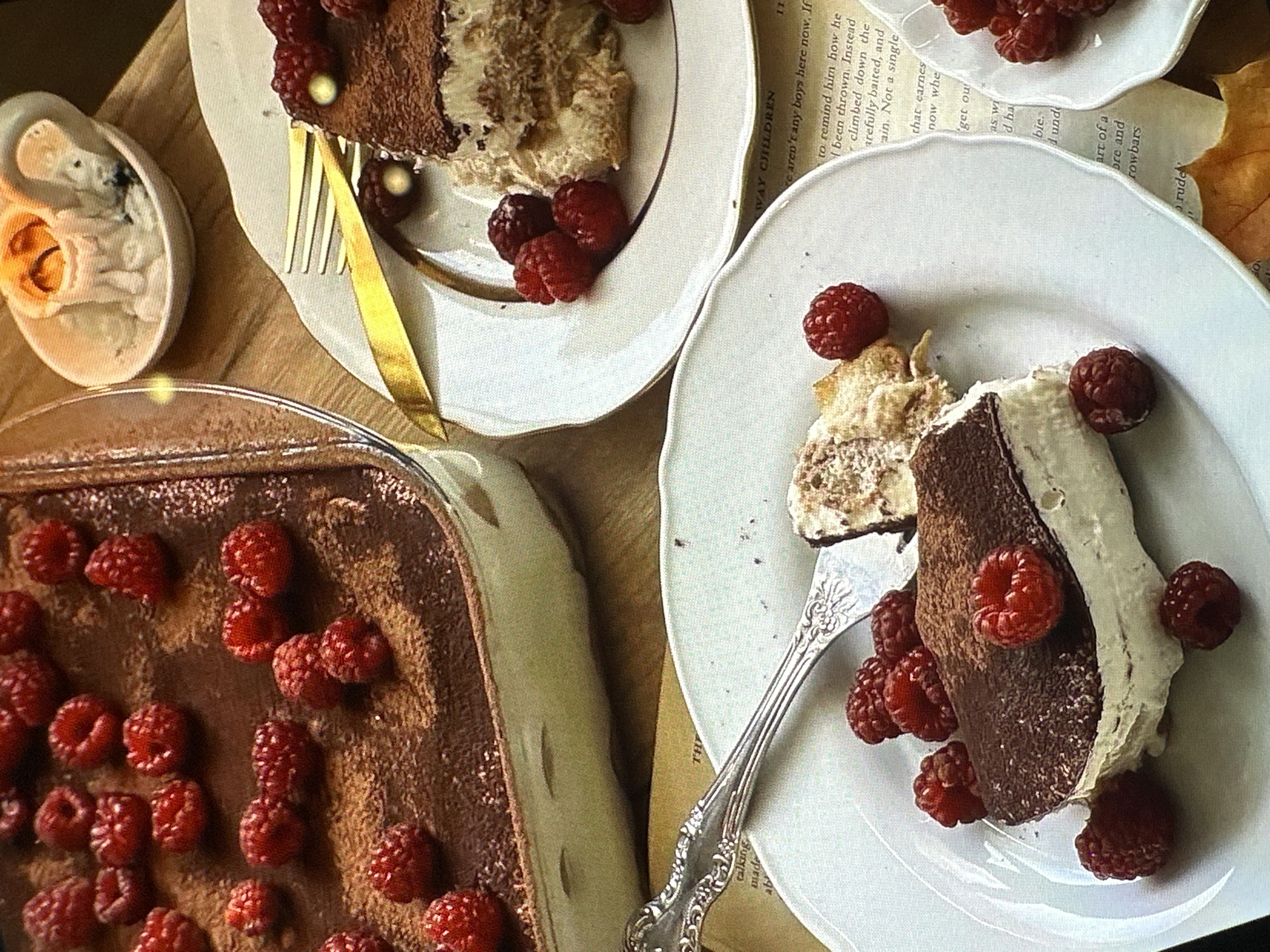 The image shows two plates of tiramisu with raspberries, positioned on what seems to be a wooden surface with an open book underneath. A dish of tiramisu covered with raspberries sits to the left. Utensils are present: a gold-colored fork on the top plate and a silver fork on the bottom plate. A small decorative object, possibly a candle holder, is to the left of the dish of tiramisu. There is no indication that this image was taken in Yerevan, Armenia.