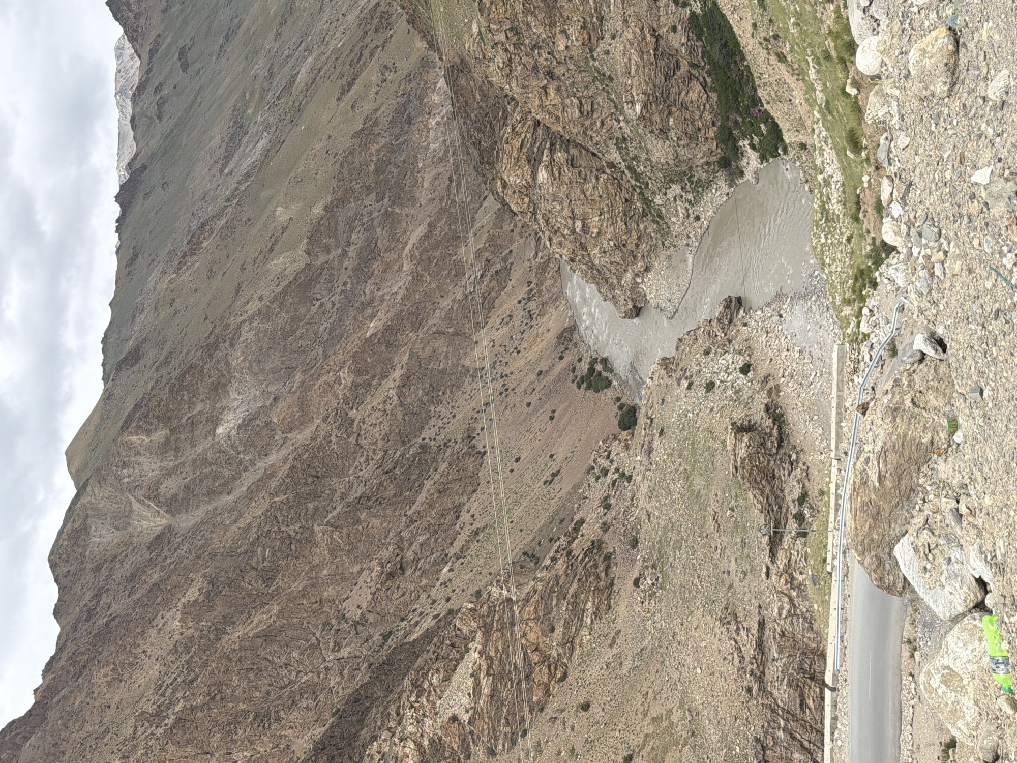 An overcast day in Reru, India, showcases a majestic, rugged mountain valley. A wide, muddy river flows through the center, carving its path between steep, rocky slopes. The mountains, predominantly brown and tan, are textured with fissures and scree, with sparse patches of dry grass clinging to the inclines. In the foreground, a paved road runs along the edge of the valley, bordered by a metal guardrail and scattered rocks. A bright green plastic bottle lies on the ground near the road, alongside other debris. Power lines are strung across the valley, suspended between poles on the mountainsides. The overall scene is one of raw, natural beauty, a testament to the powerful forces of erosion and geological formation.