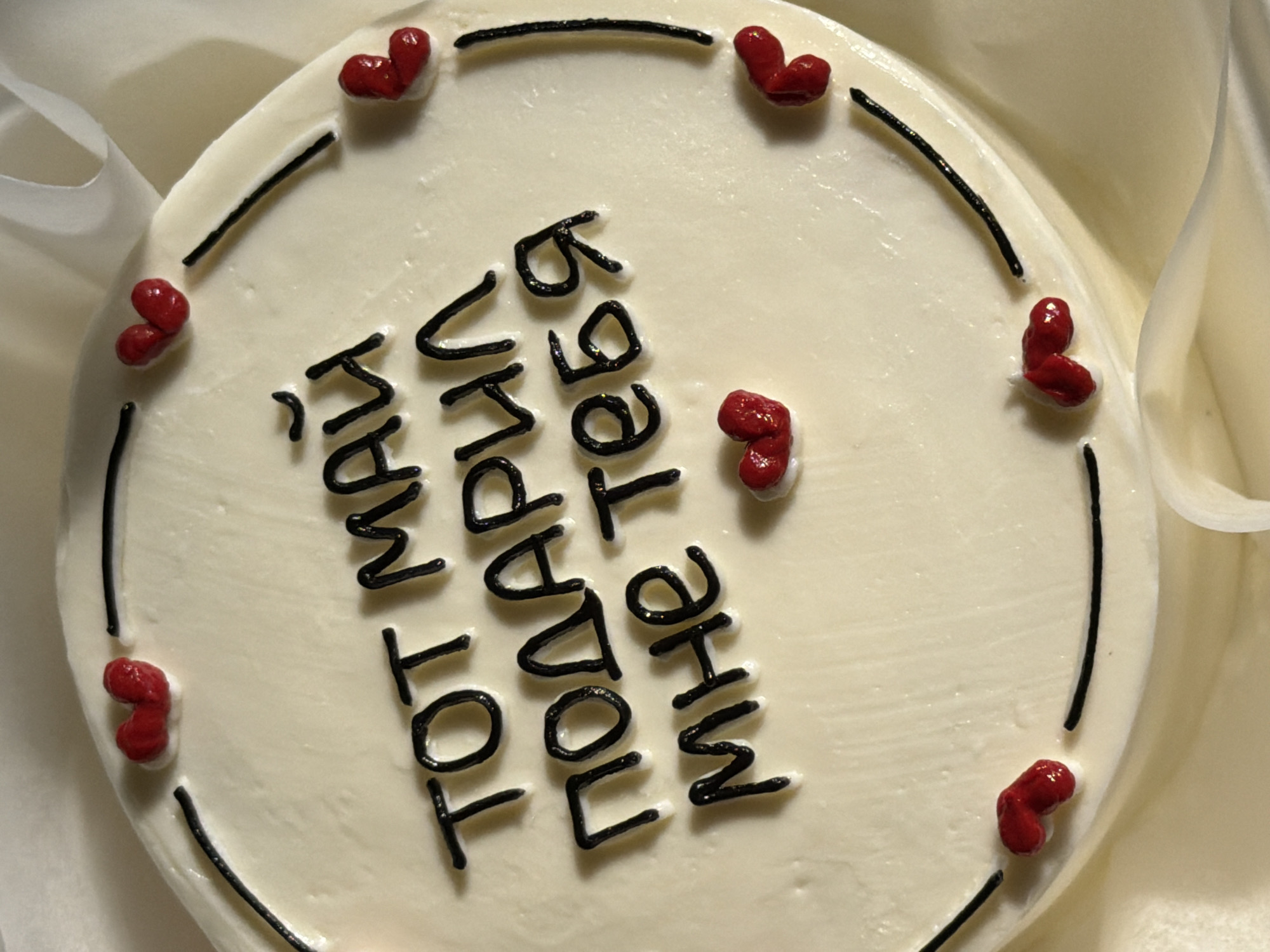 A top-down, close-up shot of a round cake, likely vanilla-flavored, adorned with a simple cream-colored frosting. Around the cake's perimeter, there is a thin, dark line of icing punctuated by small, red heart-shaped decorations. In the center of the cake, lettering written in dark icing spells out a message. The cake is set against a backdrop of pale, off-white fabric, possibly a tablecloth or wrapping paper. The image does not depict any individuals or a location in Tel-Aviv, Israel.