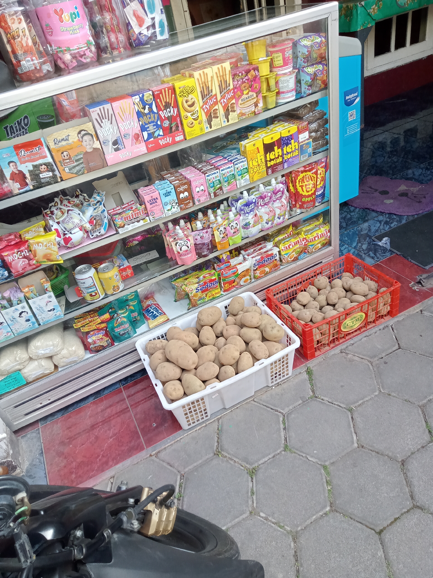 A street-side stall in Batu, Indonesia, displays an assortment of packaged snacks and produce. A glass-fronted refrigerator case is stocked with various candy bars, biscuits, and drinks. On shelves visible through the glass, there are brands like Pocky, Yupi, and Indomilk. Outside the case, two large white plastic crates are filled with potatoes. A black motorcycle is parked in the foreground, partially obscuring the view of the stall and the paved ground. The stall appears to be set up on a sidewalk with hexagonal paving stones.