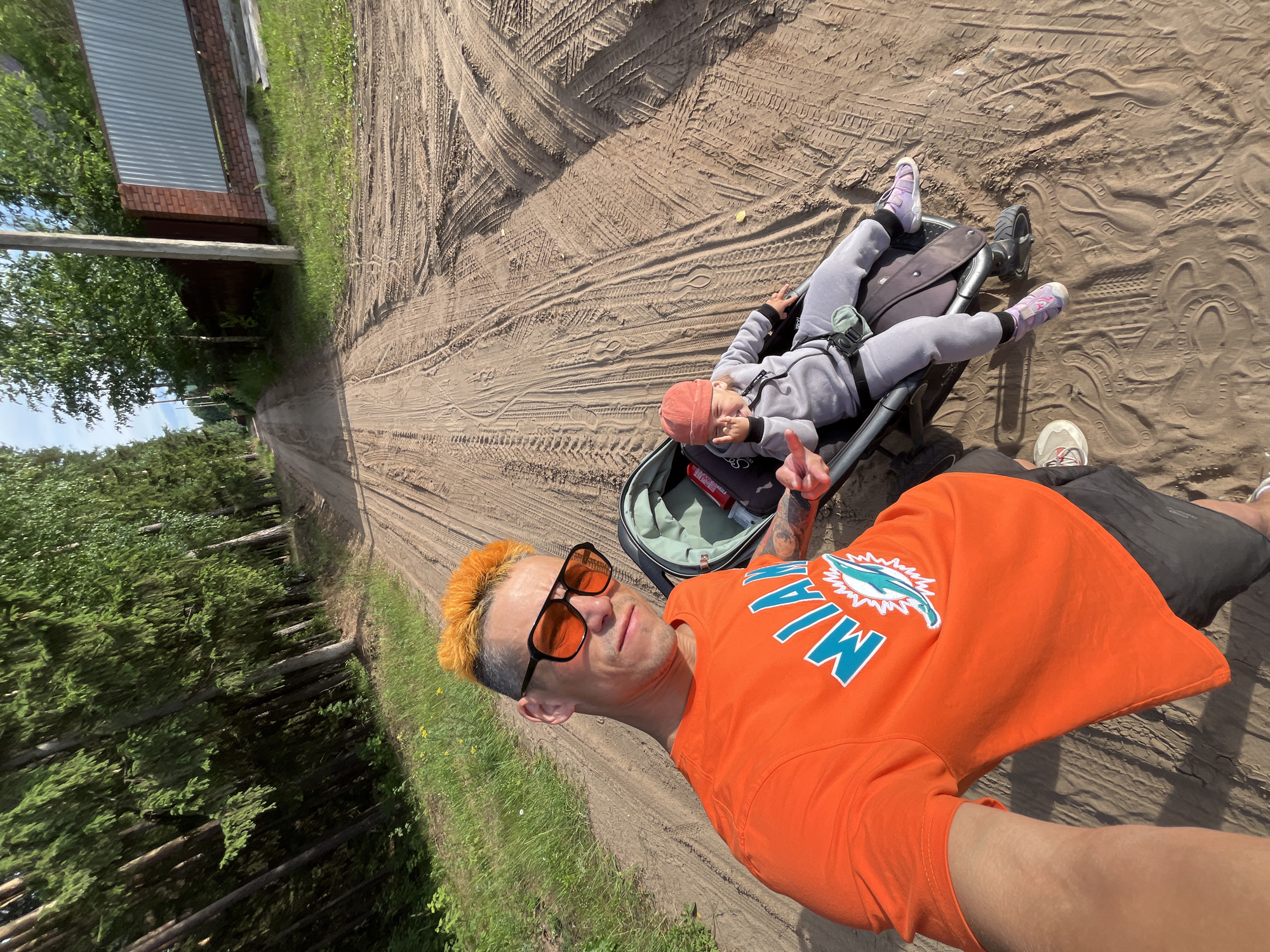This media file features two people in an outdoor, rural setting. In the foreground, a man with bright orange hair and sunglasses is taking a selfie. He is wearing an orange t-shirt with a "Miami" graphic in blue and white. His left arm is visible, showing tattoos. He appears relaxed, with a slight smile.
Behind him, a child is lying in a stroller. The child is wearing a grey tracksuit and a pink beanie. The child is looking towards the camera and holding up two fingers in a peace sign. The stroller is dark grey with a green interior.
The scene is a dirt road with visible tire tracks. To the left of the road, there is a dense forest of pine trees and a grassy area. To the right of the road, there is a building with a corrugated metal roof and brick accents, suggesting a rural structure. The sun is shining brightly, indicating it is daytime, and the weather appears to be warm. The overall mood is casual and family-oriented, likely a leisurely outing in the countryside. The location context provided is Chaikovsky, Russia.
