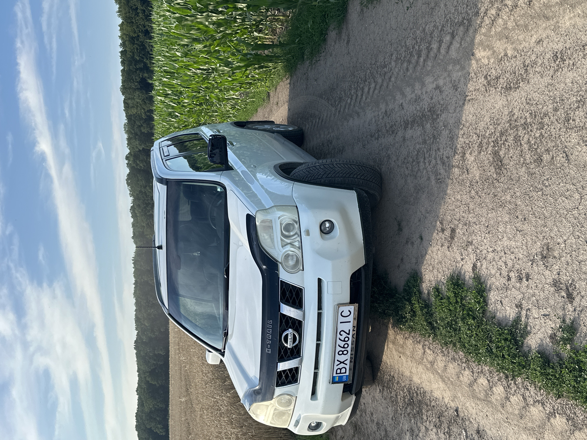 A white Nissan X-Trail is parked on a dirt road in a rural area. The car is facing the camera, with its front bumper and grille clearly visible. The license plate reads "BX 8662 IC". To the left of the car is a field of ripe wheat, and to the right is a field of tall green corn. In the background, a dense line of trees forms the horizon under a partly cloudy blue sky. The scene appears to be located in Slobidka-Zalisetska, Ukraine. There are no people or animals visible in the image. The overall environment suggests a sunny day in the countryside.