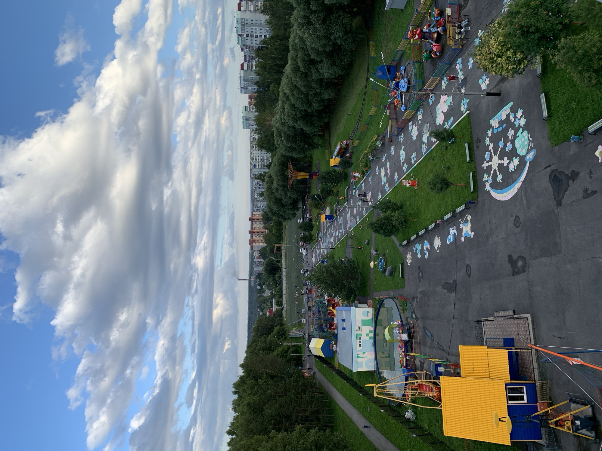 This is an aerial, eye-level photograph of a public park and playground in Prokopyevsk, Russia. The park is situated in a residential area, with apartment buildings visible in the background. The playground features a variety of attractions, including swings, a small carousel, a Ferris wheel, and a kiddie train ride. There are also several wading pools, some with decorative swan boats. The ground of the playground is paved with asphalt, and there are colorful painted designs on the pavement, including flowers and cartoon characters. Green grass areas with trees and benches are interspersed throughout the park. A soccer field is visible in the middle ground. The sky is partly cloudy, with large white clouds against a blue sky. Several people are present in the park, including children playing on the equipment and adults walking around. The overall impression is one of a well-maintained and active community park.