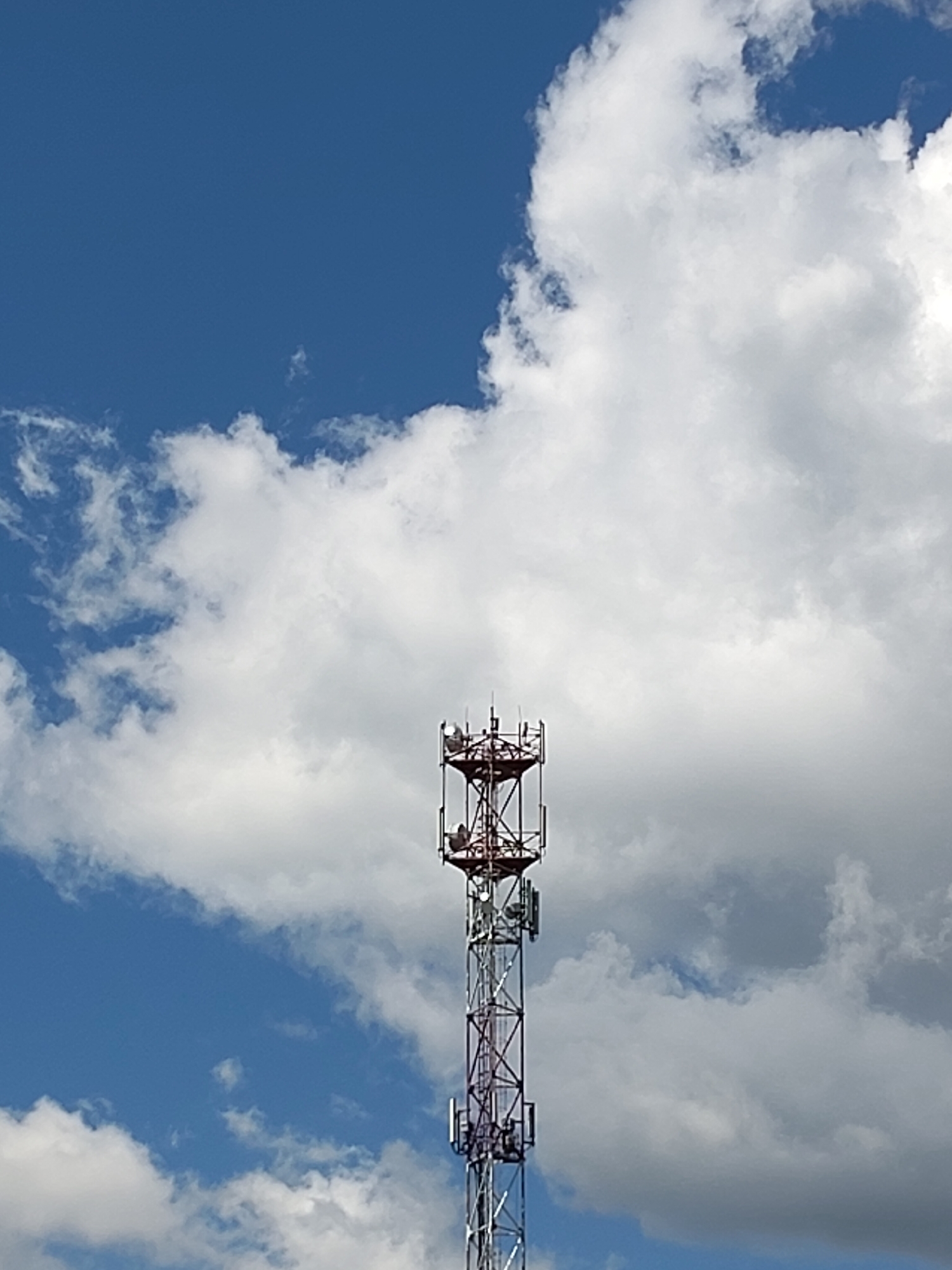 The image shows a tall telecommunications tower set against a bright blue sky filled with fluffy white clouds. The tower is a lattice structure with a red-painted upper section and a silver-painted lower section. Several antennas and equipment boxes are visible attached to the tower. The sky suggests it is daytime, likely with fair weather, as indicated by the clear blue sky and bright white clouds. The scene is outdoors, and the presence of a telecommunications tower implies a setting that requires connectivity, possibly in a semi-urban or rural area. No people are visible in the image. There is no visible text in the image. The location context provided is Ходаево, Russia.