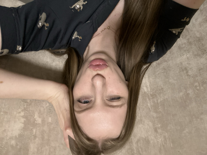 An indoor, close-up photograph features a female subject positioned against a textured, light brown wall. The specific location within Chaikovsky, Russia, is not identifiable from the visual content. The subject has long, light brown hair, which falls over her shoulders. Her left eye is closed in a wink, and her lips are pursed in a forward "duck face" gesture. Her right eye is partially open, directed towards the right side of the frame. Her left hand is raised, with the palm resting against the top-rear of her head. She is wearing a dark-colored (likely navy or black) collared short-sleeved shirt patterned with multiple light-colored sloth figures, some appearing to hold cups. A delicate, gold-toned chain necklace with a small circular pendant is visible on her neck.