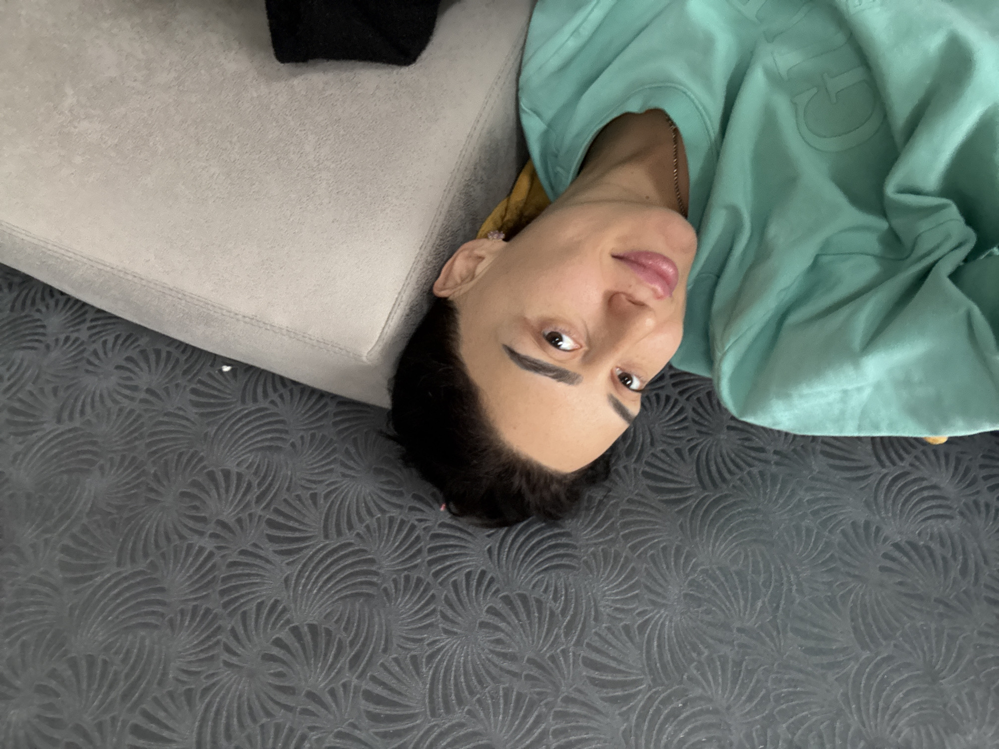 A person is depicted lying down indoors in Tbilisi, Georgia, looking directly at the camera. The image is oriented with the person's head positioned towards the lower right. The individual, who appears to be female, has dark, short hair and a neutral to slightly pleasant expression with slightly parted lips. They are wearing a teal or mint green long-sleeved top, possibly a sweatshirt, which has an embossed design featuring a large, prominent "G" and other indistinct letters. A thin, gold-colored chain necklace is visible around their neck, and a small, light-colored earring is in their left ear.
The person's head rests near a light gray upholstered sofa or cushion, which occupies the upper left portion of the frame. The dominant surface in the image is a dark gray carpet or rug, featuring a repeating pattern of intricate, fan-like or shell-like motifs. The indoor lighting appears soft, without harsh shadows. A dark, indistinct object is visible in the far upper left corner, possibly another part of the sofa or an item of clothing.