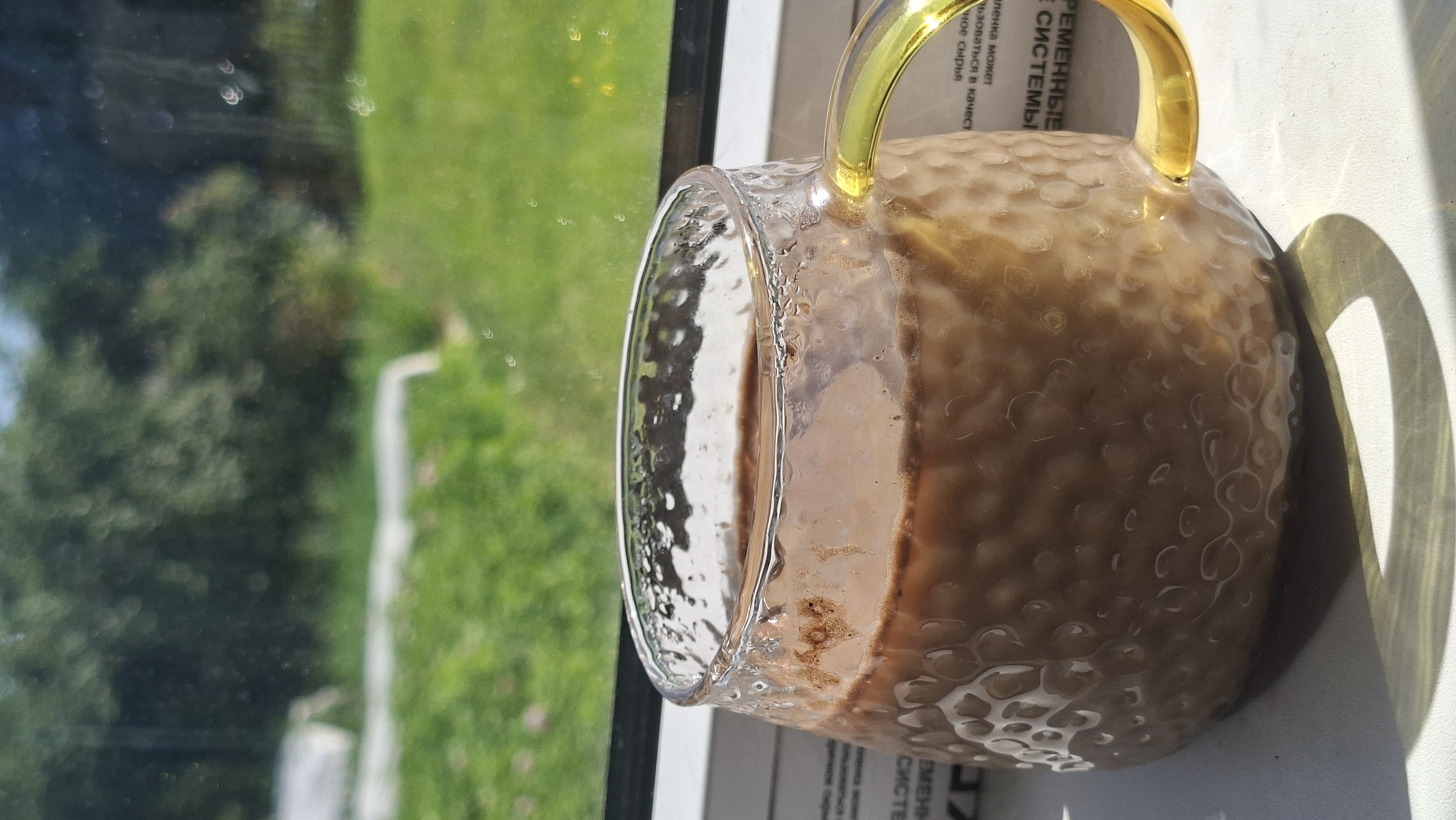A close-up shot shows a glass mug filled with a light brown beverage, possibly coffee or hot chocolate, sitting on a white surface. The mug has a textured, hammered glass design and a bright yellow handle. The beverage has a frothy layer on top and appears to be warm, with condensation visible on the glass. In the background, through a window, lush green grass and trees are visible, suggesting an outdoor setting. The sunlight casts a distinct shadow of the mug onto the surface. The scene is set near a window, with some printed text visible beneath the mug on a white paper or book. The location is identified as Забалоцкі сельскі Савет, Belarus.