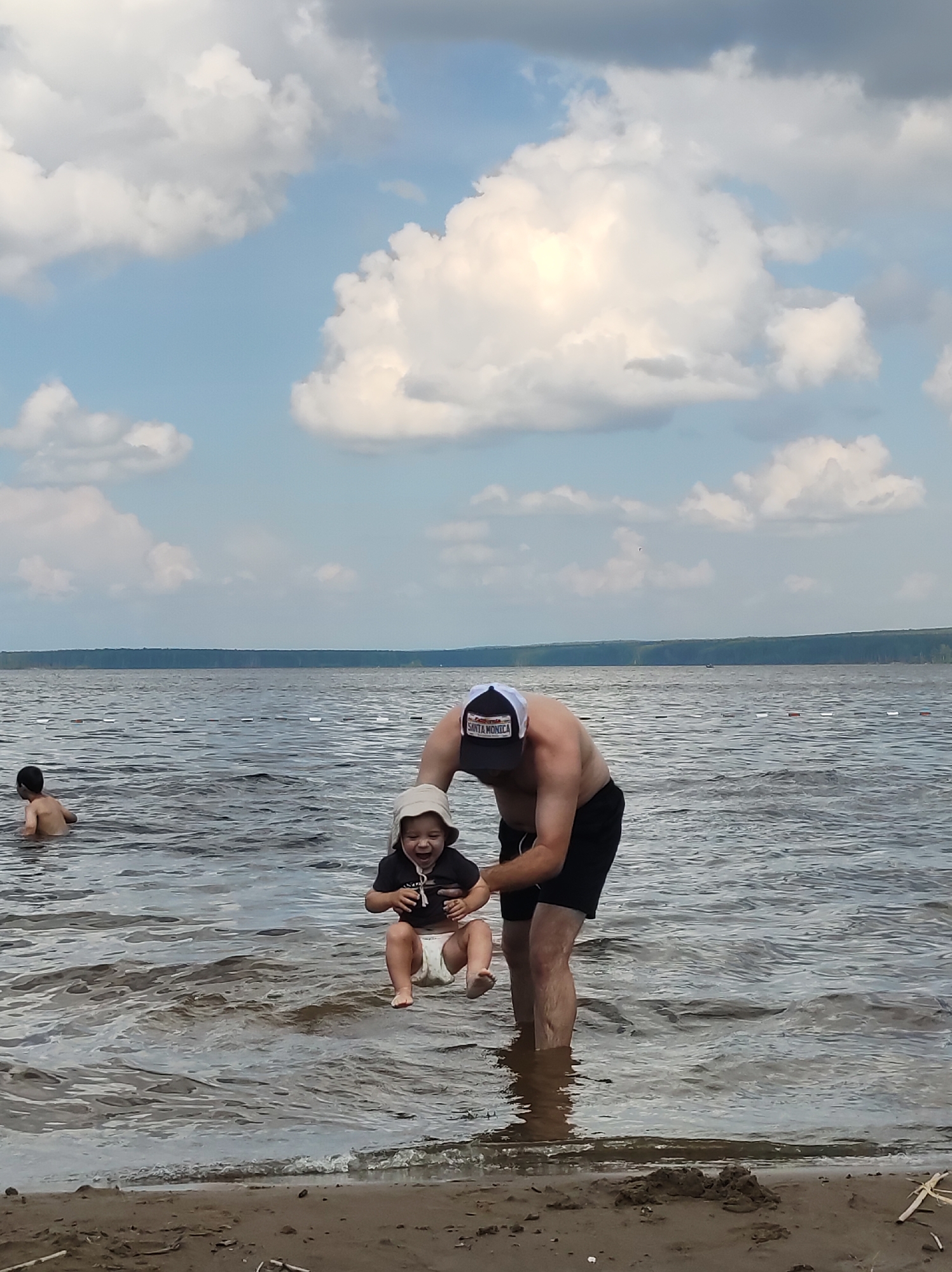 A man is holding a baby in the shallow water of a lake. The man is wearing a baseball cap and black shorts. The baby is wearing a diaper and a hat. In the background, another child is swimming in the lake. The sky is blue with white clouds. The scene appears to be a recreational area, possibly a beach or park, located in the city of Chaikovsky, Russia, known for its water bodies.