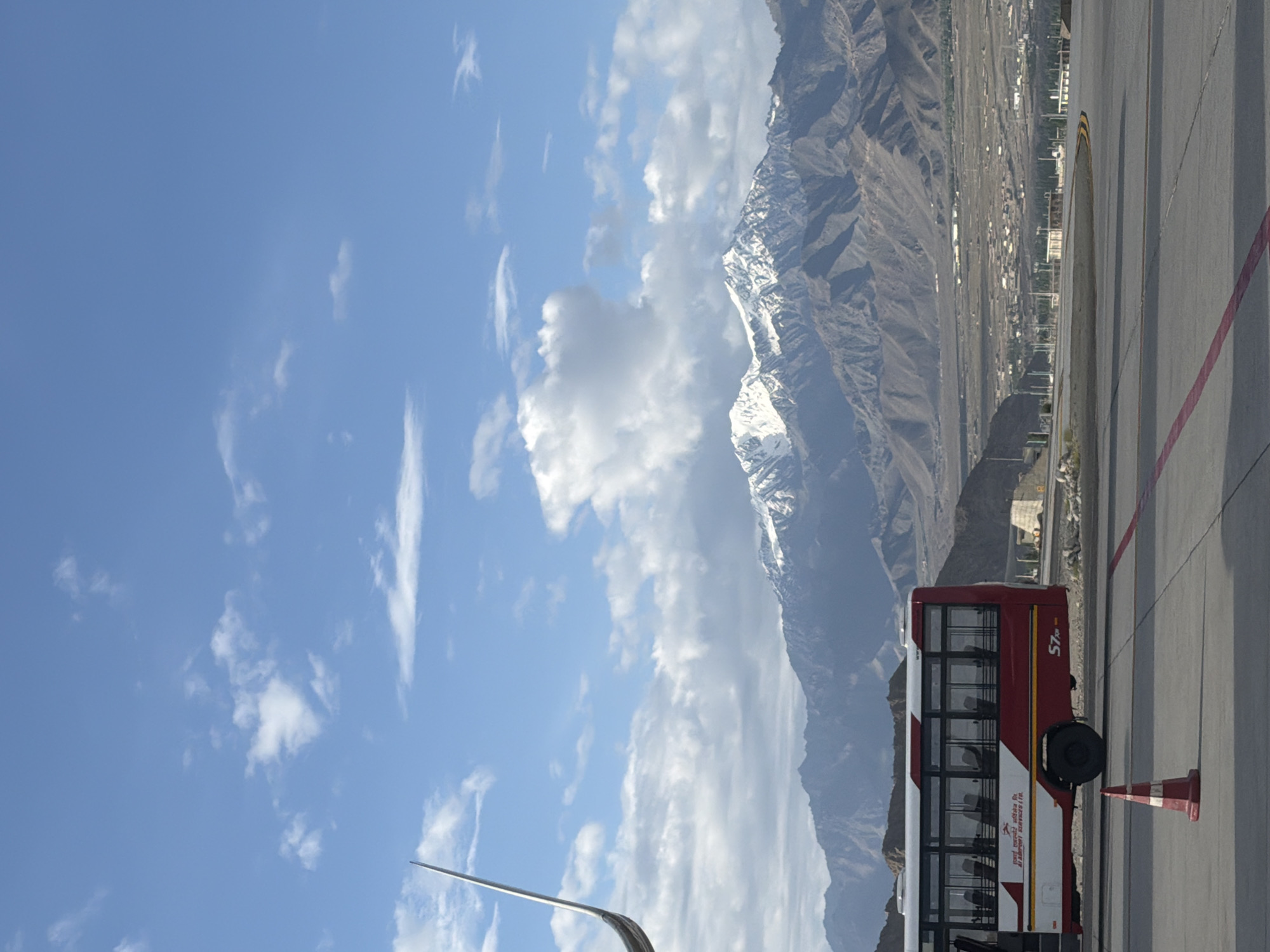 In Leh, India, a red and white bus with tinted windows is parked on an airport tarmac. The bus has "AI AIRPORT SERVICES LTD." written on its side, and "S7" is visible near the rear wheel. A red and white traffic cone is placed in front of the bus. In the background, a vast, arid landscape stretches out, leading to a mountain range with snow-capped peaks under a bright blue sky dotted with white clouds. A large, silver airplane wing is visible in the upper left corner of the image, suggesting the bus is at an airport.