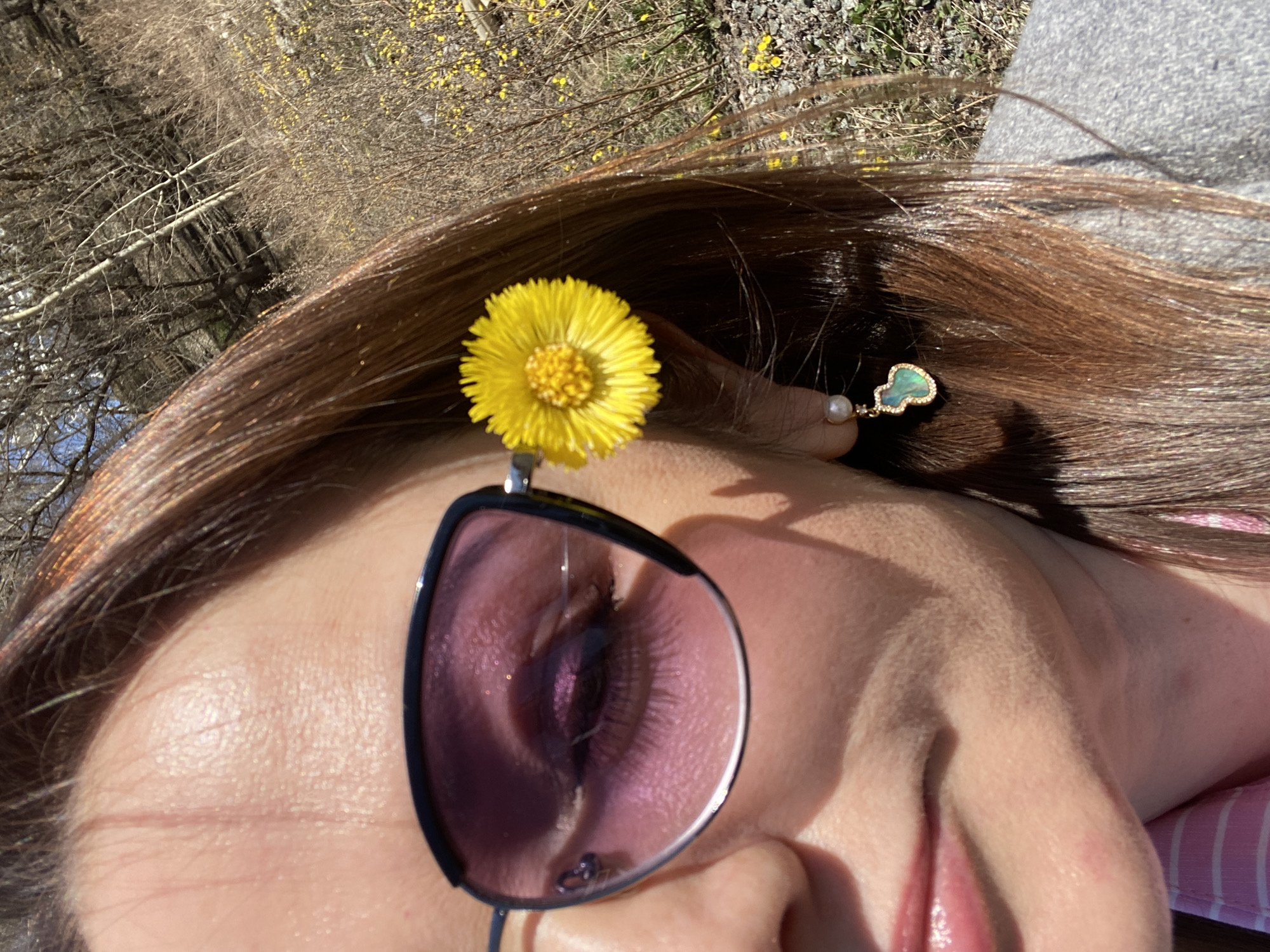 A close-up, tilted view features a woman's face, partially obscured by sunglasses with dark purple lenses. A bright yellow flower is perched on the frame of the sunglasses above her right eye. She is wearing an earring with a pearl and a green, kidney-shaped pendant. The woman has brown hair. The background shows bare branches and other yellow flowers. The location of Yekaterinburg, Russia is not supported by the given information.