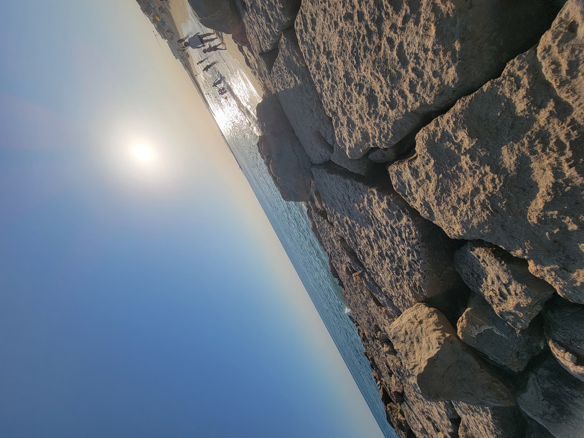 The image depicts a sunny day at a beach, likely in Quarteira, Portugal, as suggested by the presence of the Quarteira skyline in the background. The foreground is dominated by large, rough, light-colored rocks, likely part of a breakwater or natural rock formation. These rocks are positioned at an upward angle from the lower left to the upper right of the frame.
Beyond the rocks, the calm, blue ocean stretches out, meeting a sandy beach. Several people are visible on the beach and in the shallow water, appearing to be engaged in recreational activities such as swimming or walking. The sun is bright and high in the sky, casting a strong glare on the water's surface. The overall atmosphere is one of a typical summer day at the seaside.