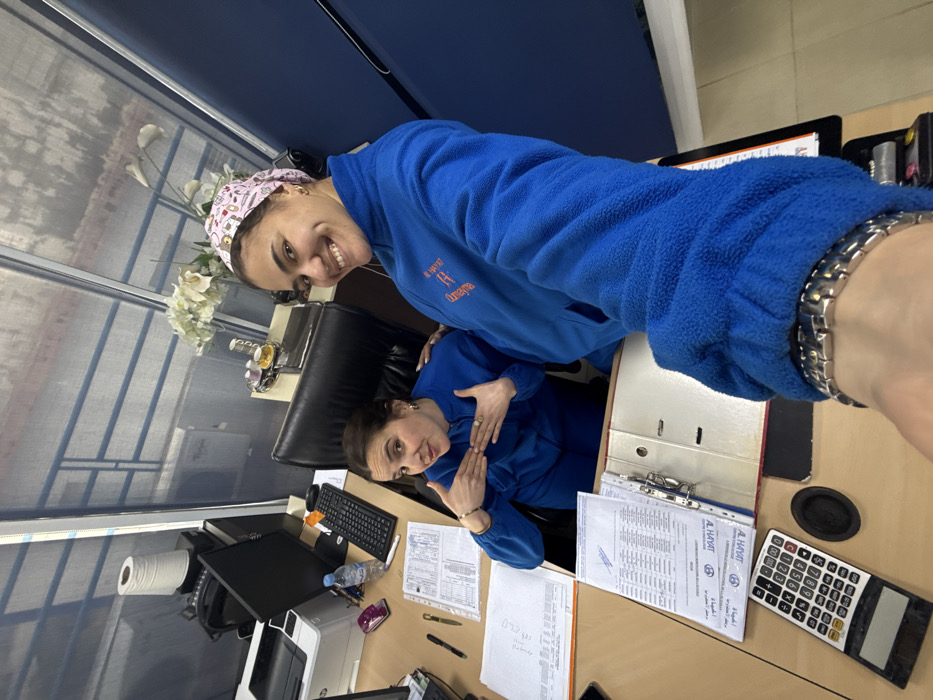 Two women pose for a photo at a desk. The woman in front wears a blue jacket and a patterned cap. The woman behind her also wears a blue jacket and makes a hand gesture. The desk is cluttered with a keyboard, documents, a calculator, and office supplies. There is a window and blinds to the left, with flowers visible near the window.