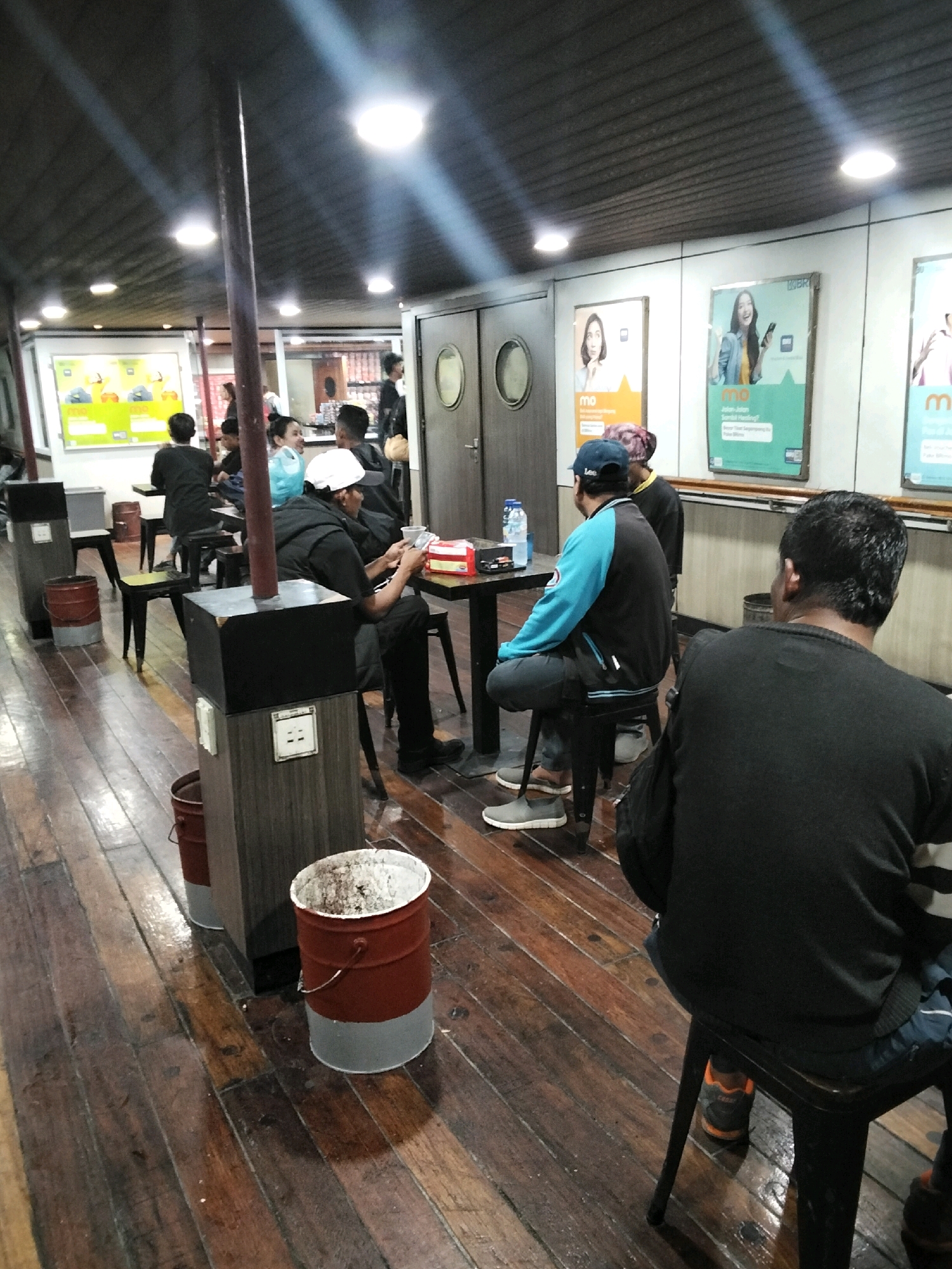 The image shows an indoor scene with several people sitting at tables and on chairs. The environment appears to be a casual dining or waiting area with wooden flooring and a dark ceiling with overhead lighting. Several posters are visible on a wall in the background, featuring advertisements with women.
In the foreground, on the right, a person wearing a black sweater is seated on a stool, facing away from the camera. To the left, another person wearing a blue and black jacket and a blue cap is seated on a stool, looking down at a table. A man in a white cap and black clothing is sitting at the same table, holding what appears to be a phone or small device.
Further back, several other individuals are seated at tables. One person is wearing a black t-shirt and is looking towards a table. There are also tables and chairs set up, with some drink bottles and containers on them.
The overall impression is a relaxed atmosphere in a dimly lit indoor space. The mention of Batam, Indonesia, suggests this scene might be within a terminal, a restaurant, or a similar public venue in that city.