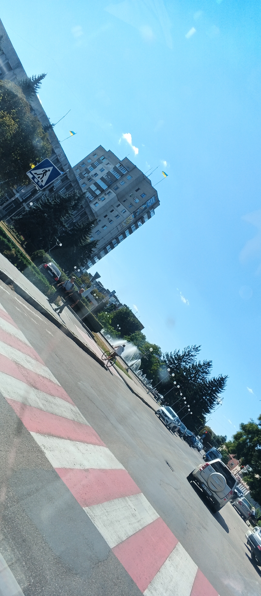 The image depicts a street scene in Smila, Ukraine, viewed from an angle that suggests it was taken from a vehicle. The perspective is tilted, with the sky occupying the upper right portion of the frame. A multi-story, light-colored apartment building with numerous windows and balconies dominates the background. Two Ukrainian flags are visible flying from the roof of this building. In the foreground, a pedestrian crossing with red and white stripes is prominent. A person is visible riding a bicycle near a fountain with multiple water jets. Several cars are parked along the street and driving on the road. Trees and a pedestrian crossing sign are also present in the scene. The overall environment appears to be an urban setting on a clear day.