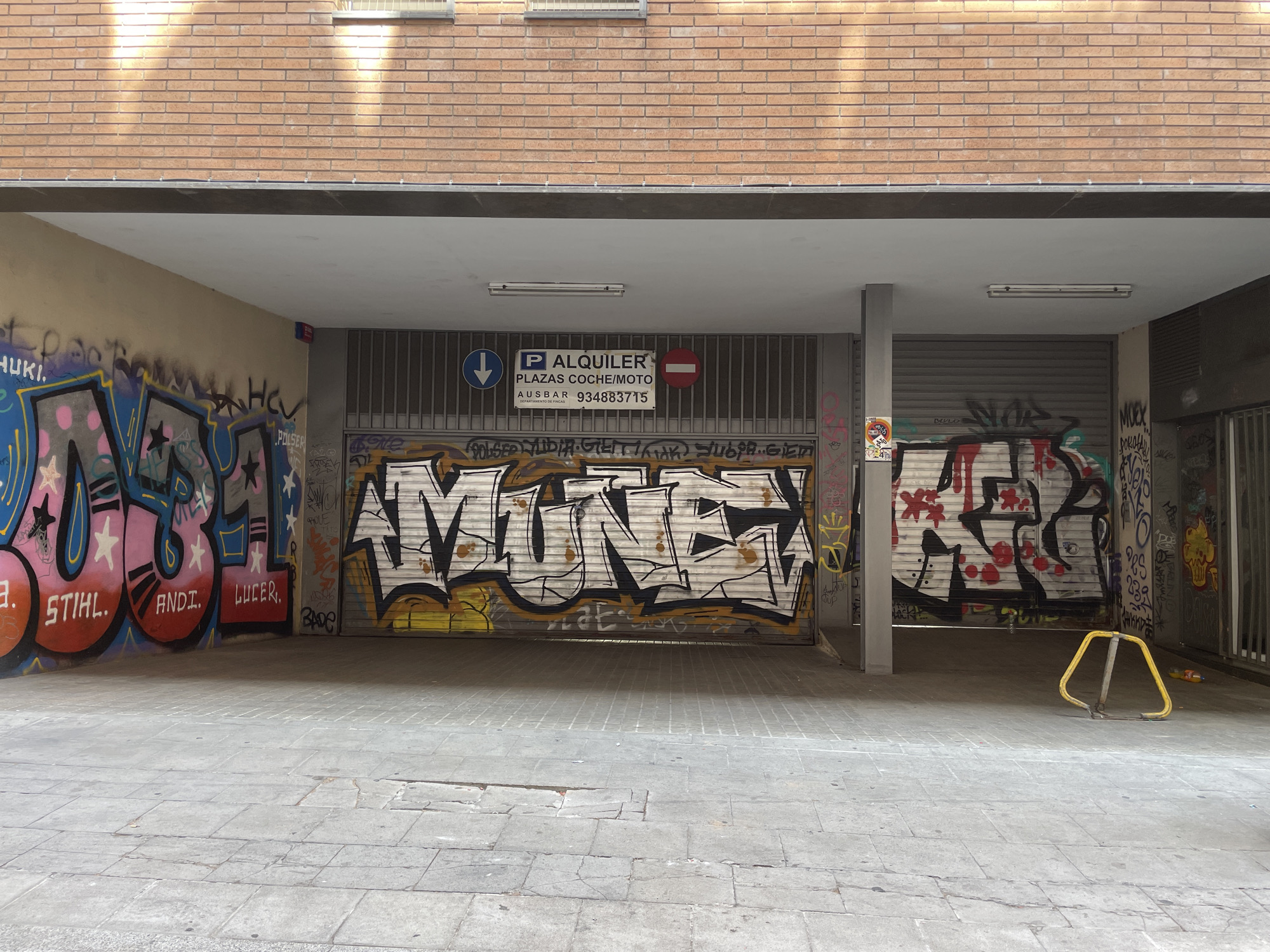 The image captures a street scene in Barcelona, Spain, likely an underpass or covered walkway. The scene is dominated by graffiti art covering several metal rolling doors and parts of the walls. In the center, a large white graffiti piece with black outlines and yellow accents reads "MUNFA". To the left, another graffiti tag in pink, blue, and black is visible. To the right, a red and white graffiti piece with black outlines is present. Above the central rolling door, a sign indicates "P ALQUILER PLAZAS COCHE/MOTO AUSBAR 934883715", suggesting a parking rental service. The ground is paved with gray square tiles. A yellow metal barrier is situated in the lower right portion of the image. The overhead structure is a brick wall with two horizontal windows. The lighting suggests it is daytime, with sunlight casting shadows.