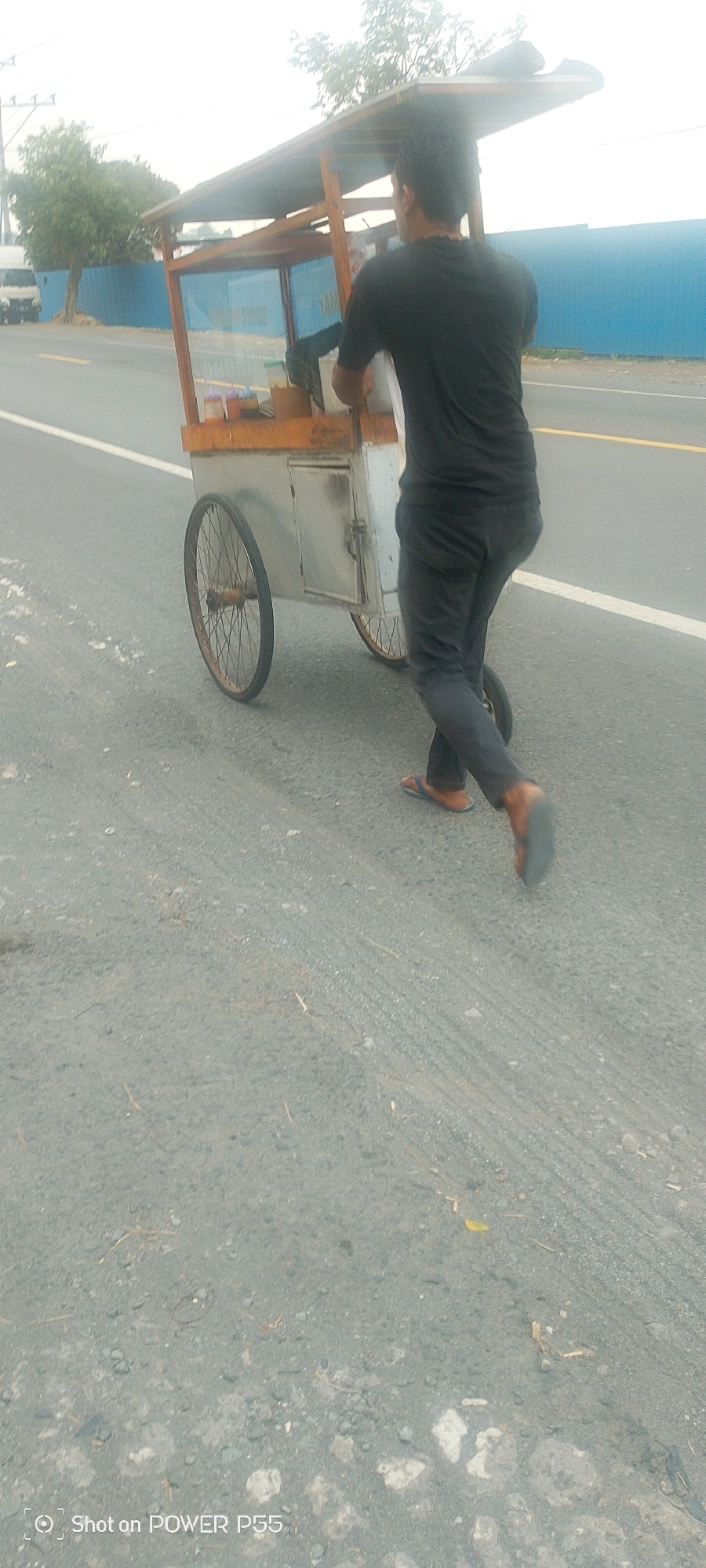 A person wearing a black t-shirt and dark pants pushes a food cart on a paved road. The food cart has a wooden frame with a metal enclosure and wheels. The person is walking away from the camera, pushing the cart forward. The background shows a blue fence, trees, and a white vehicle. The road has yellow and white lines marking lanes. The lighting suggests it is daytime.