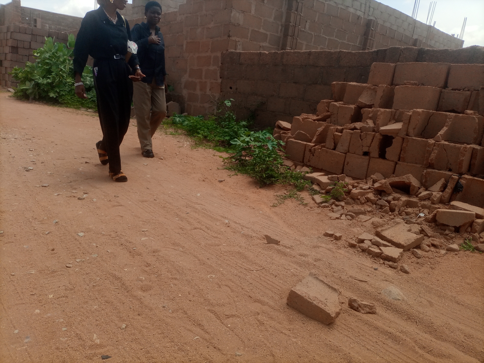 Two individuals are walking on a dirt road in a developing urban area that appears to be the city of Tumfure, Nigeria. The person on the left, dressed in black, is a woman wearing earrings, a necklace, a bracelet, black trousers, and brown sandals. She is walking with her left hand extended. The person on the right, also dressed in black, is a man wearing a dark shirt and light brown trousers, with his right hand on his chest. The environment consists of unfinished brick buildings in the background, piles of broken bricks, and scattered vegetation. The ground is covered in reddish-brown dirt with visible tire tracks and small stones. The sky is overcast.