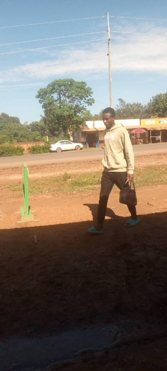 The image shows a young man walking on a dirt road in Kivaywa, Kenya (0.56, 34.806). He is wearing a light brown hoodie, black pants, and blue flip-flops. He is carrying a small, dark brown handbag. There is a white car parked on the road behind him. The man is walking away from a tall wooden pole with multiple electric wires attached. There are trees and foliage in the background. The ground is dirt, with some grass and foliage growing in areas. The sky is clear and blue. The image is taken from a slightly elevated angle.