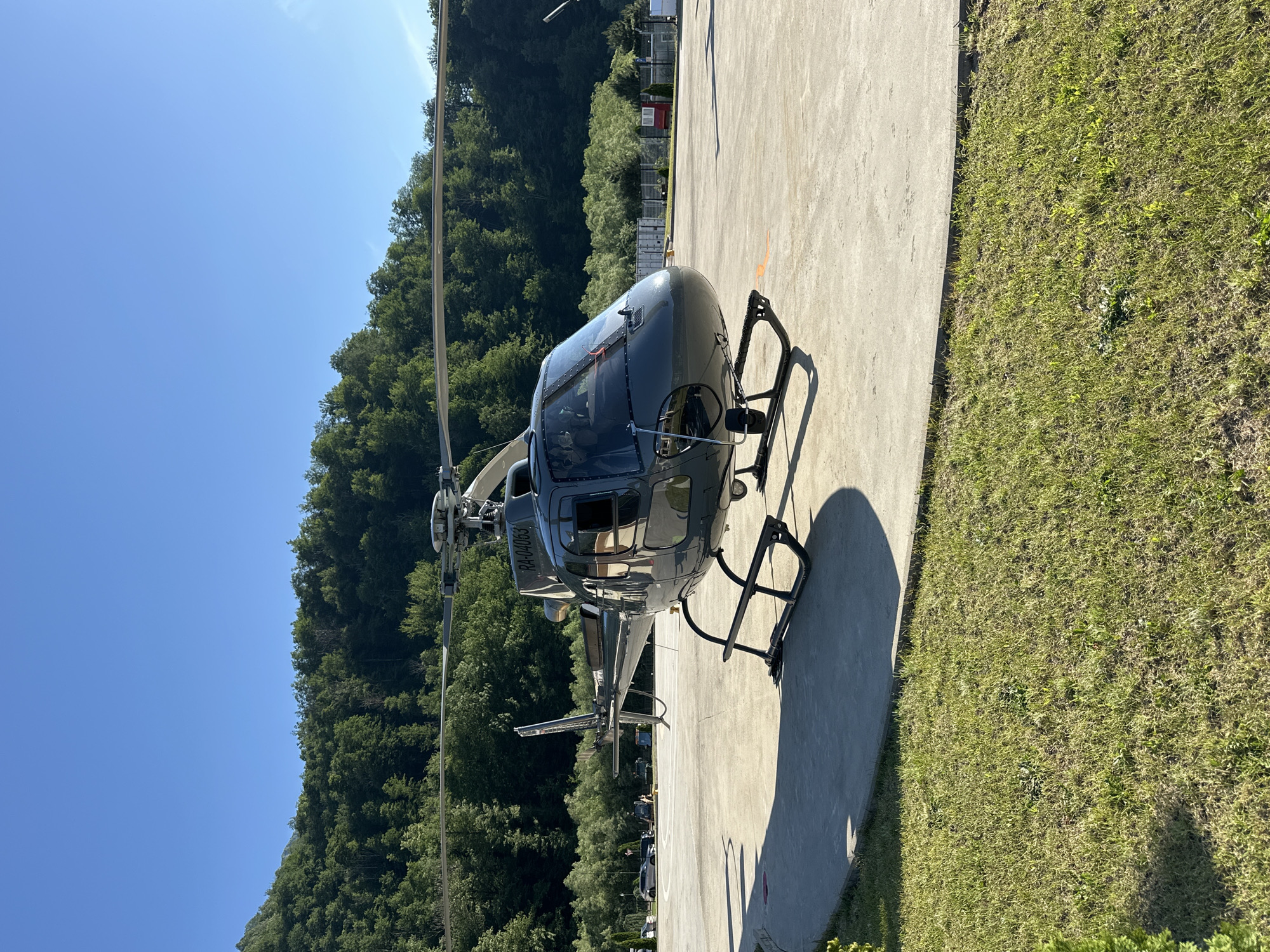 A dark gray helicopter is parked on a concrete helipad. The helicopter has a single main rotor and a tail rotor, with a visible registration number RA-04063 on its tail. The cockpit windows are tinted, and the landing gear is visible.
The scene is set against a backdrop of lush green trees that form a dense forest covering a hillside. The sky is a clear, bright blue, indicating a sunny day. The time of day appears to be late morning or early afternoon, given the strong shadows cast by the helicopter and the bright sunlight.
To the right of the helipad, there is a strip of green grass. In the background, beyond the helicopter and trees, several small buildings and shipping containers are visible, suggesting a small industrial or operational area. The overall impression is one of a remote, possibly mountainous, location, consistent with the context of Krasnaya Polyana, Russia. No people are visible in the image. The scene is peaceful and still, with no activity immediately apparent. The weather is clear and warm.