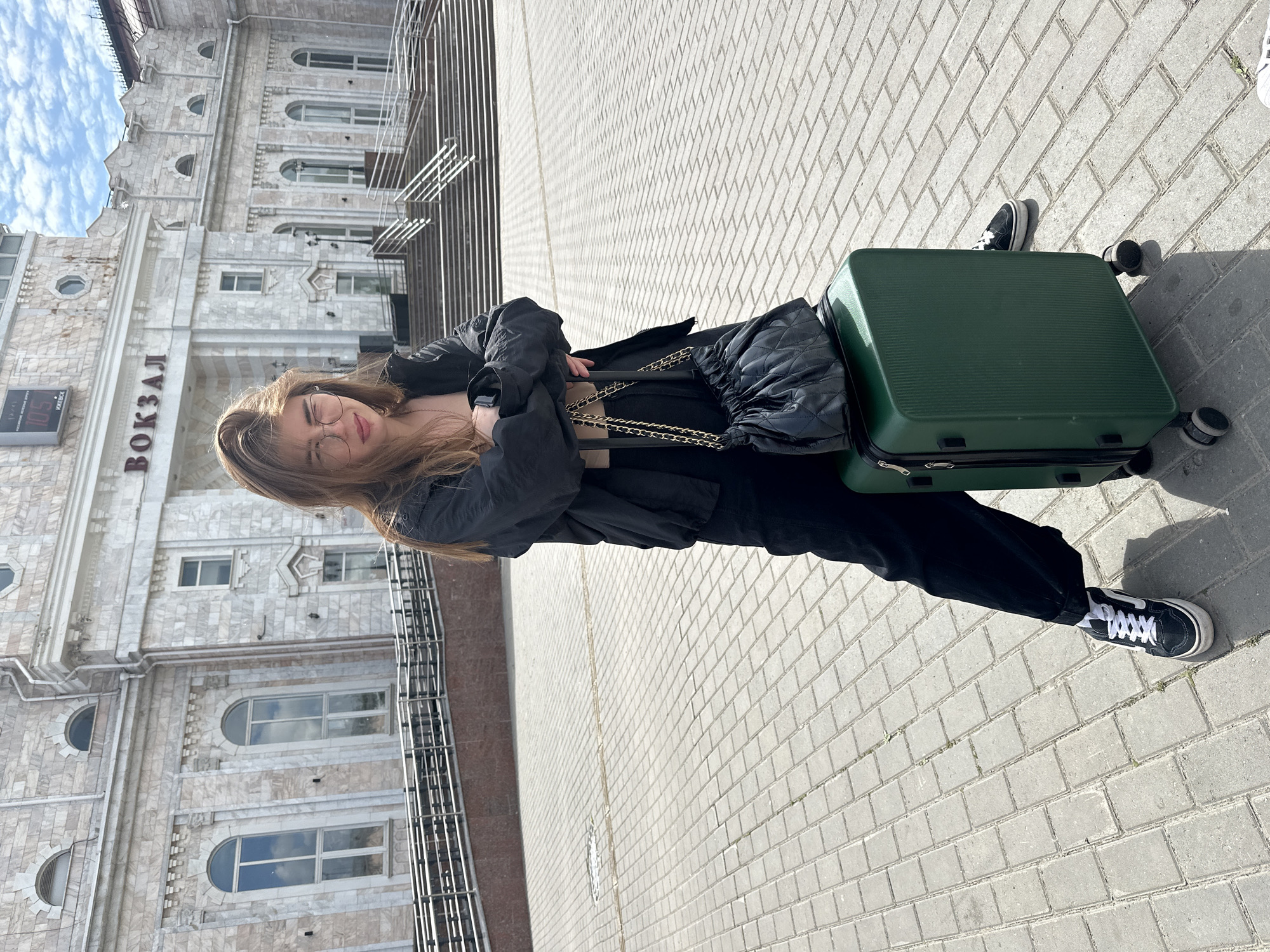 The image captures a young adult female standing on a paved outdoor area in front of a grand stone building. The building is identified as "ВОКЗАЛ" (Station) in Izhevsk, Russia, indicated by signage above the entrance and text on a digital clock display showing "ИЖЕВСК" and "10:51". The female subject has long brown hair, wears glasses, a black lightweight jacket over a light-colored top, black wide-leg trousers, and black sneakers with white laces. Her arms are crossed, and she is positioned with a dark green, rigid-shell suitcase to her right. A black quilted shoulder bag with a gold chain strap rests on top of the suitcase. The ground is paved with rectangular light grey stones. The station building in the background features an ornate facade with multiple arched windows reflecting a partially cloudy blue sky. Access to the building entrance is provided by a wide set of stairs with metal railings and a contiguous long, dark red ramp with matching railings. The scene is illuminated by bright daylight, casting distinct shadows to the right of the subject and the suitcase.