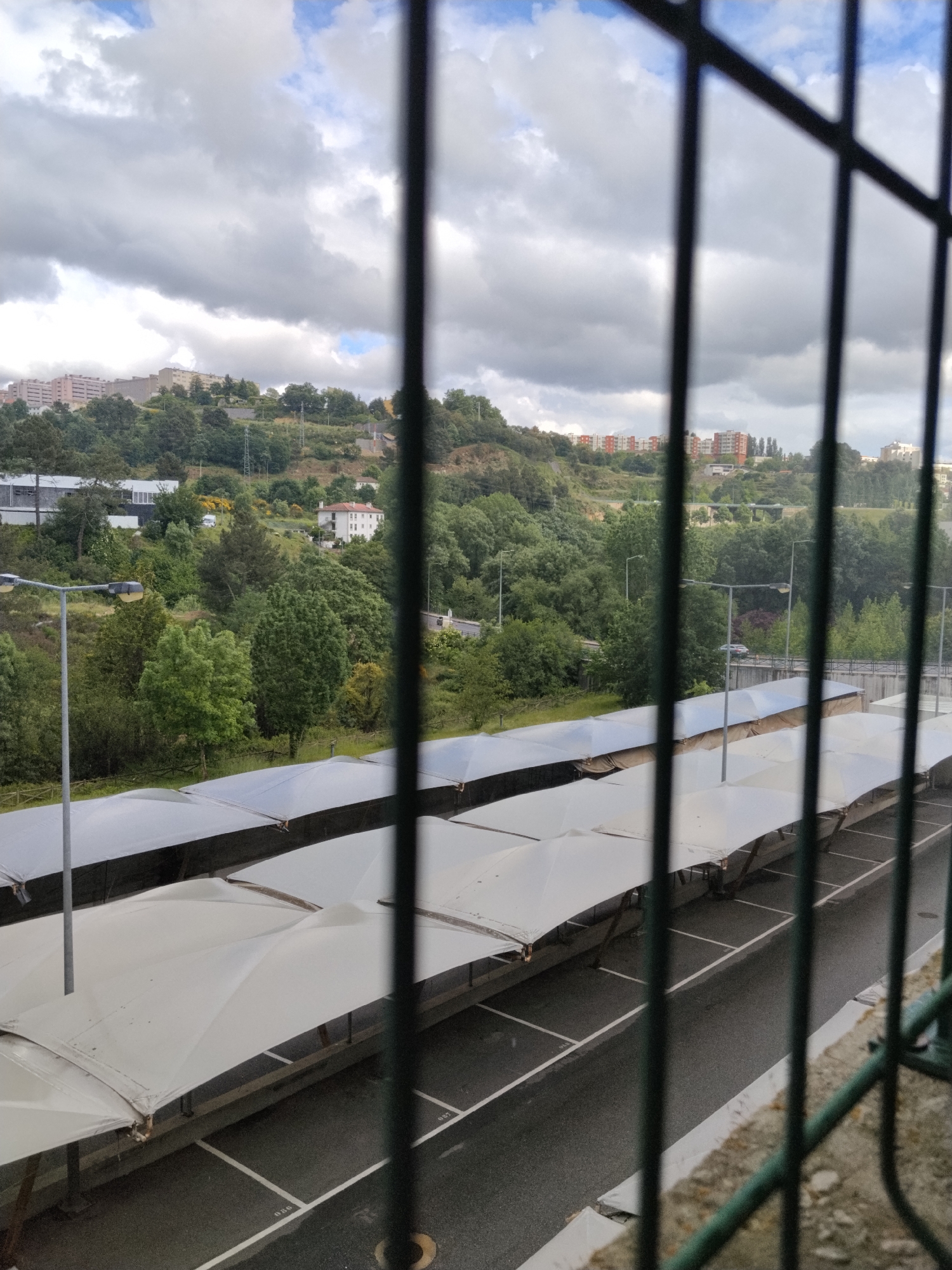 A daytime shot through a metal barred window shows a covered parking lot in Vila Real, Portugal. The parking lot features multiple rows of awning-covered spaces. Behind the parking lot, a hill rises, densely vegetated with trees and interspersed with lampposts. Buildings are visible on the hilltop, some appearing as apartment blocks. The sky is overcast with patches of blue. The focus is on the scene outside the window.
