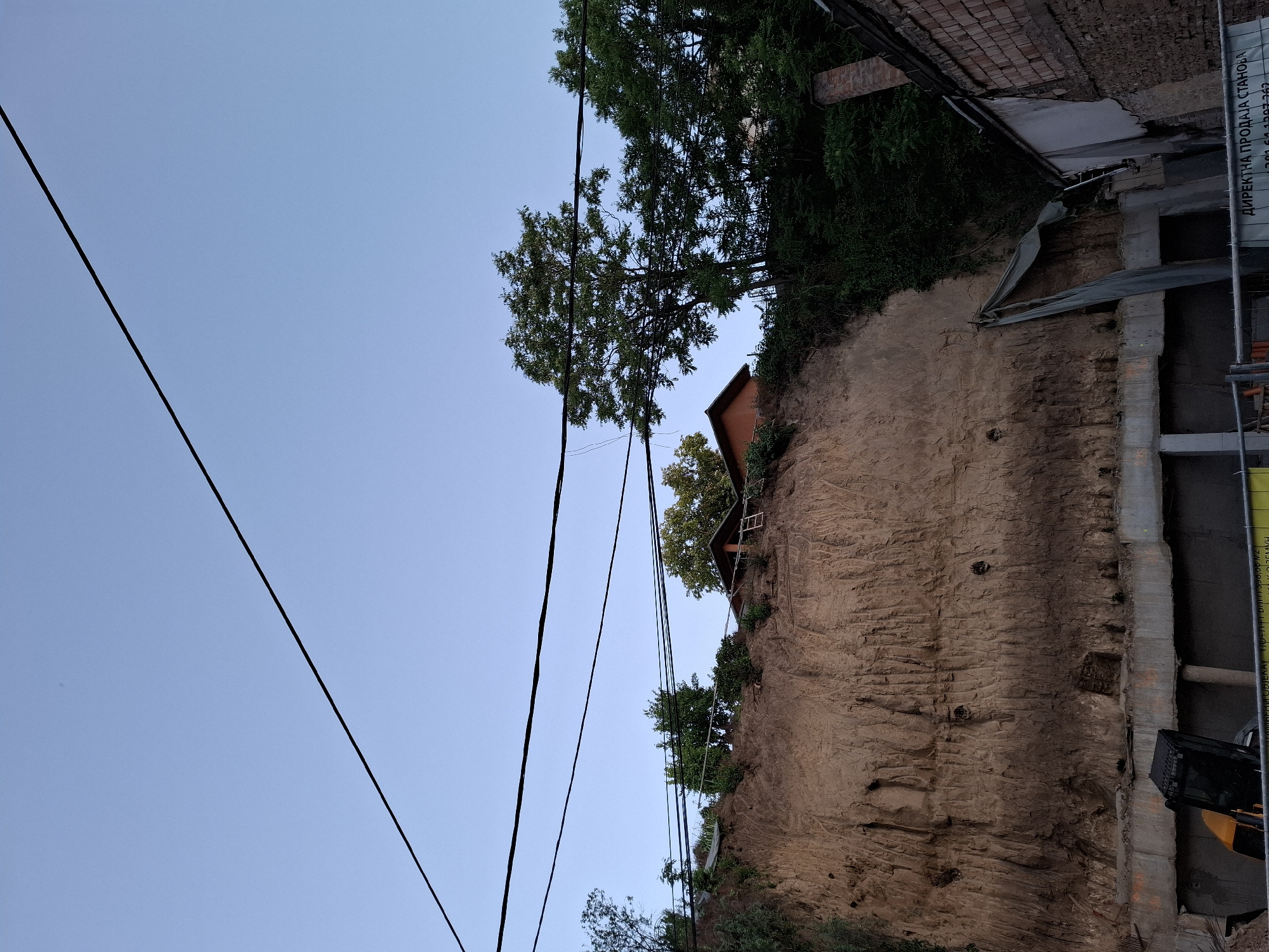 The image shows a cliff face in Zemun Urban Municipality, Serbia. A multi-story building, in the process of construction, stands at the base of the cliff. Vegetation grows on the top of the cliff face, where a small cabin is perched. Power lines stretch across the sky on the left side of the image.