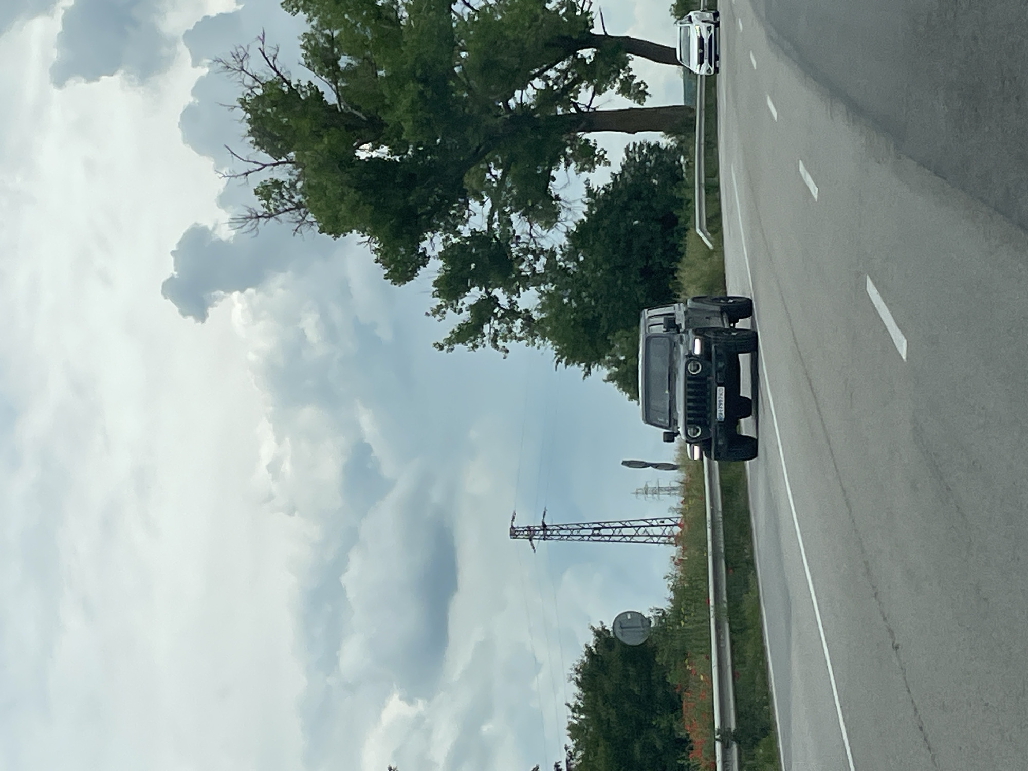 The image shows a road with two vehicles traveling on it, under a sky filled with clouds. In the foreground, a dark-colored Hummer is seen. Further down the road, a white car is also visible. The road has lane markers, and on the sides, there is greenery, including trees. In the background, there is a metal electrical transmission tower. Veselina, Bulgaria is not explicitly visible, but the scene depicts a road and environment consistent with a rural area that could be found in Veselina.
