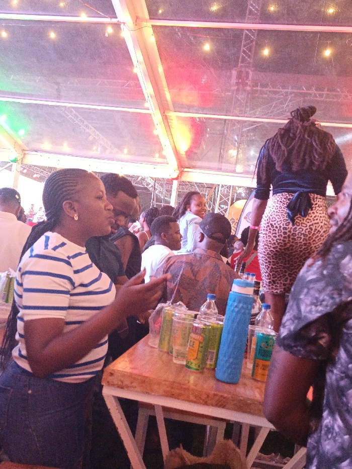 The image displays an indoor social gathering under a large translucent tent structure in Machakos, Kenya. The tent's white support beams are visible overhead, strung with small lights and illuminated by red and green colored event lighting. In the foreground, a dark-skinned female with cornrow braids, wearing a white and blue horizontally striped t-shirt and dark jeans, stands facing right with her right arm slightly raised. Adjacent to her, a light-colored wooden table with white metal legs holds multiple clear plastic cups with beverages, four green beverage cans, two clear plastic water bottles, one blue textured water bottle, and one dark bottle. To the right, the back of a dark-skinned female is visible, wearing a dark top and a leopard-print skirt, with dreadlocks or braids styled back and a pink wristband on her left wrist. The midground contains various individuals, including a male in a dark shirt and glasses, a male with a dark cap, another male in a patterned brown shirt, and a smiling female with lighter skin, among others. Background elements suggest additional event infrastructure.