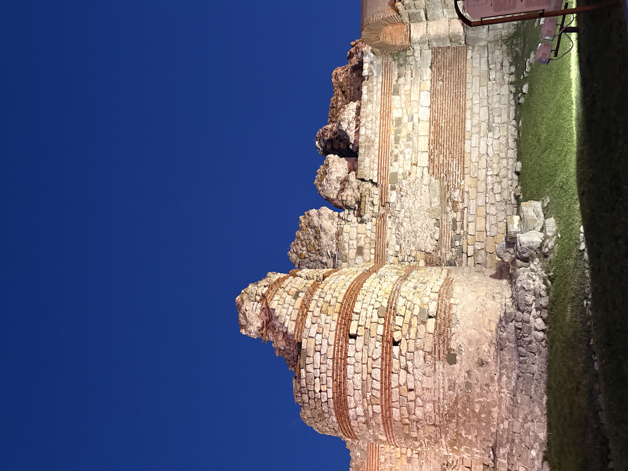The image depicts the ancient fortress walls of Nesebar, Bulgaria, under a deep blue twilight sky. The stone structure, characterized by its weathered blocks and alternating bands of red brick, shows signs of age and partial ruin. A circular tower section is prominent on the left, with sections of the wall extending to the right, featuring arched brickwork at its far end. The base of the wall is bordered by a well-maintained patch of green grass, illuminated by spotlights from the lower right of the frame. A dark, metal informational stand with a purple panel is partially visible on the right side, along with a bench and its lighting fixture. The scene is devoid of any people.